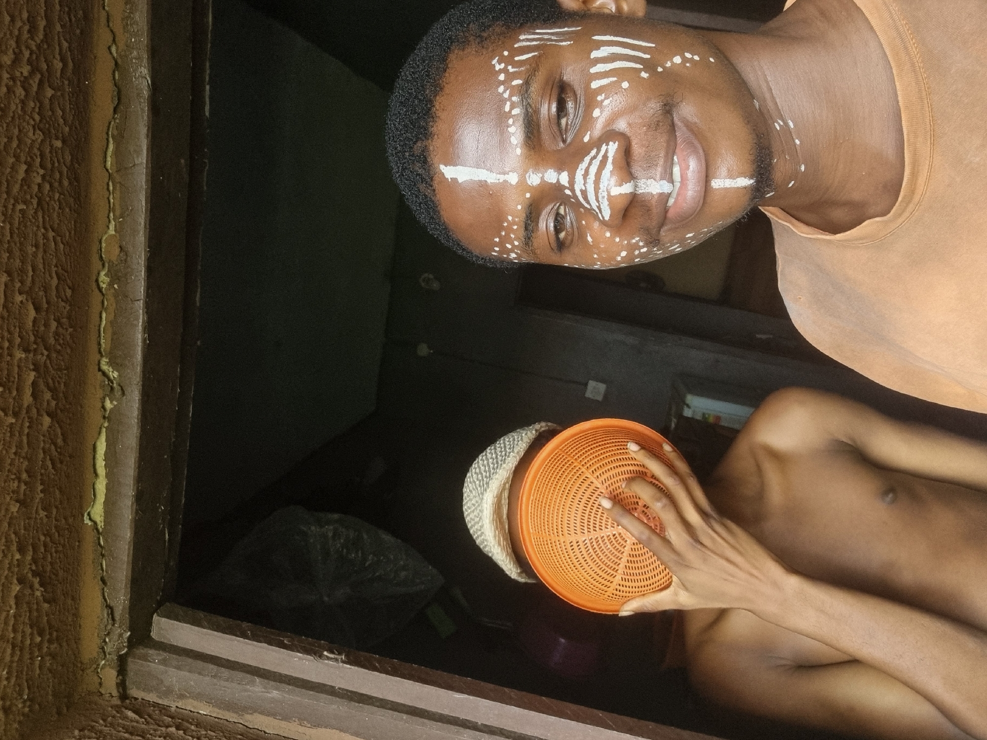 The image, taken in Nkpor, Nigeria, features two young individuals in a dimly lit indoor setting. The primary subject, a Black man, is positioned vertically, facing the camera with a gentle smile. His face is distinctly adorned with white ceremonial or ritualistic markings, including horizontal lines across his forehead and nose, and scattered dots on his cheeks and chin. He has short, dark, curly hair and a light beard, and is wearing a plain light orange t-shirt.
In the lower right portion of the frame, a second person, appearing to be a young man, is partially visible. He is shirtless, and his face is obscured by an orange plastic colander which he holds with one hand. He also wears a light-colored, woven hat.
The background is dark, indicating either night, evening, or an interior space with minimal lighting. On the far left, a textured, earthy-brown wall with some light green discoloration or growth along a seam leads to a dark wooden frame, possibly part of a window or doorway. The scene conveys a sense of cultural engagement or playful interaction, highlighted by the face paint and the obscured face of the second individual. There is no visible text in the image.