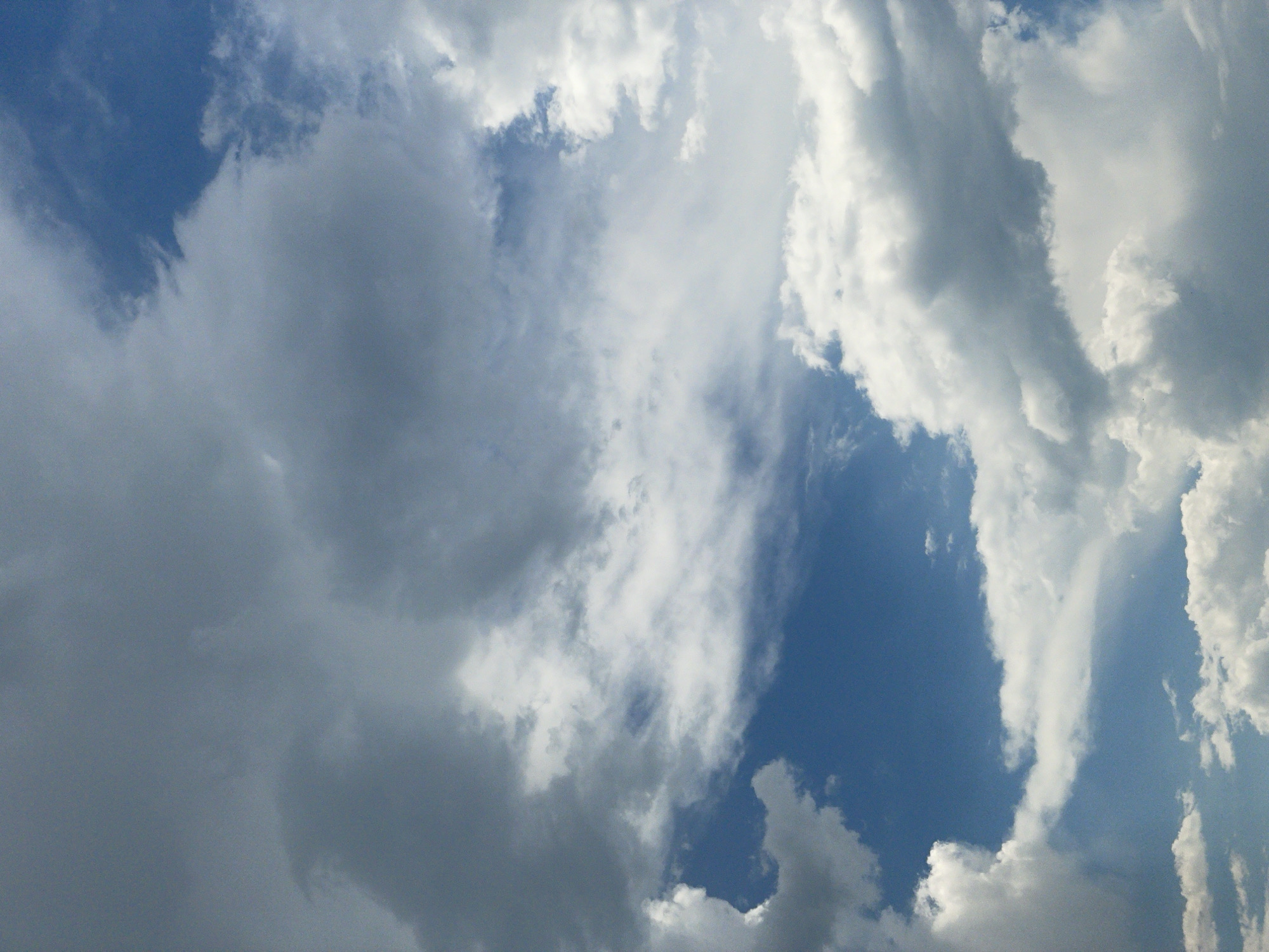 The image is a photograph of a cloudy sky taken from Vodní Újezd, Czechia. The sky is filled with a mix of dark, heavy clouds and lighter, wispy formations. Patches of blue sky are visible between the clouds. The photograph is a close-up of the sky, with no ground features or any other objects visible. The lighting suggests it is daytime, but the cloud cover is significant.
