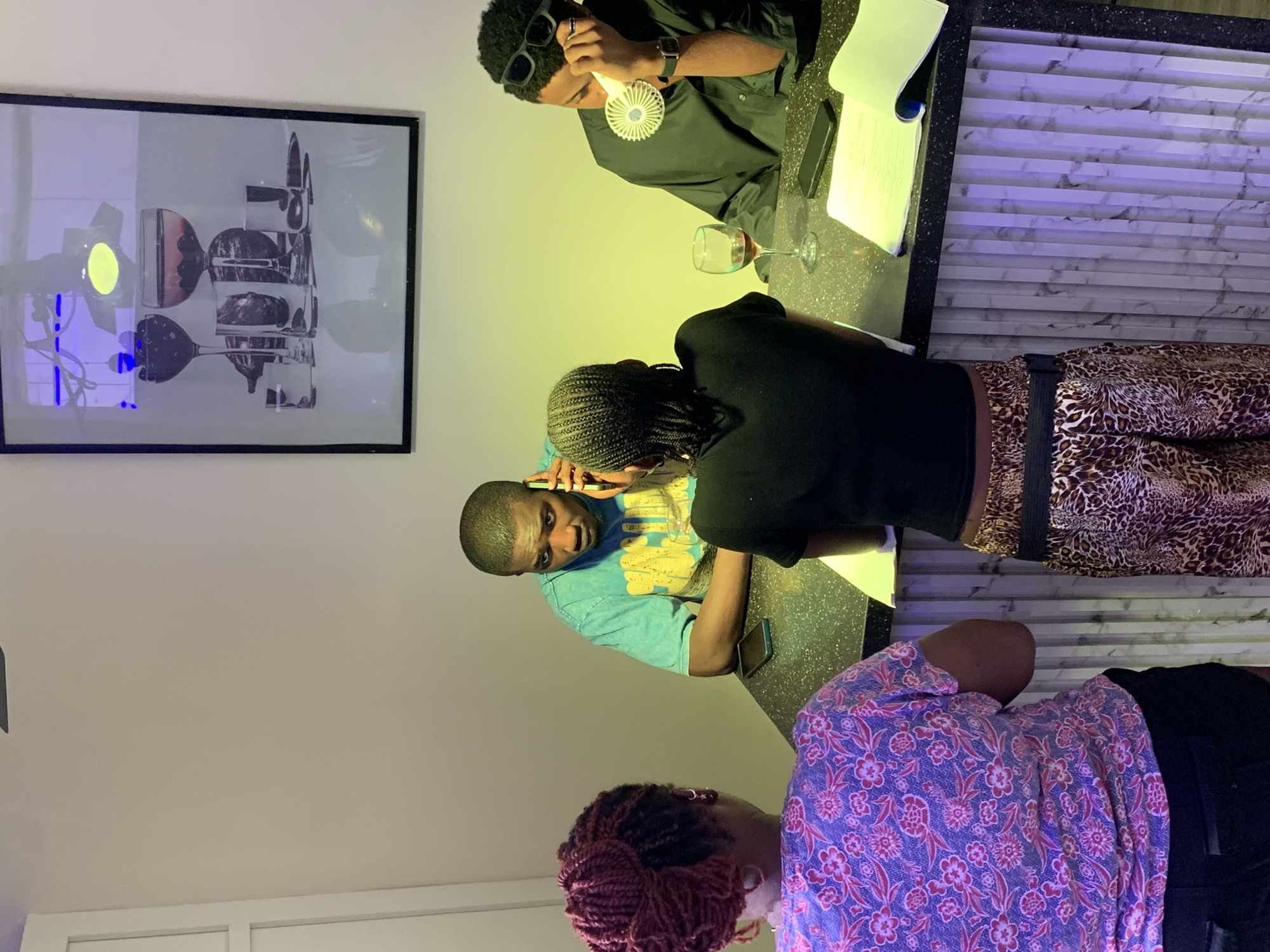 An indoor scene depicts four individuals gathered around a dark-speckled counter. On the left, a female with reddish-purple braided hair in a bun wears a floral top, her back to the camera. To her right, a second female with dark braided hair, wearing a black top and leopard-print pants, leans forward, reviewing papers on the counter. Opposite them, two males are positioned at the counter. The male in the center, wearing a teal t-shirt, holds a dark smartphone to his right ear, engaged in a conversation, with another phone resting on the counter. The male on the right, wearing a dark long-sleeved shirt and sunglasses on his head, holds a small white portable fan to his face. A wine glass containing red liquid, an open notebook, and a black smartphone are arranged on the counter in front of him. The background wall is light-colored and features a large framed picture depicting various glasses and dark fruits. The lower portion of the counter's front is paneled with a light-colored, marble-patterned material, illuminated by a subtle purple light. A yellowish-green light source illuminates the wall area directly behind the two males. The specific location within Moba, Nigeria, is not evident from the image.