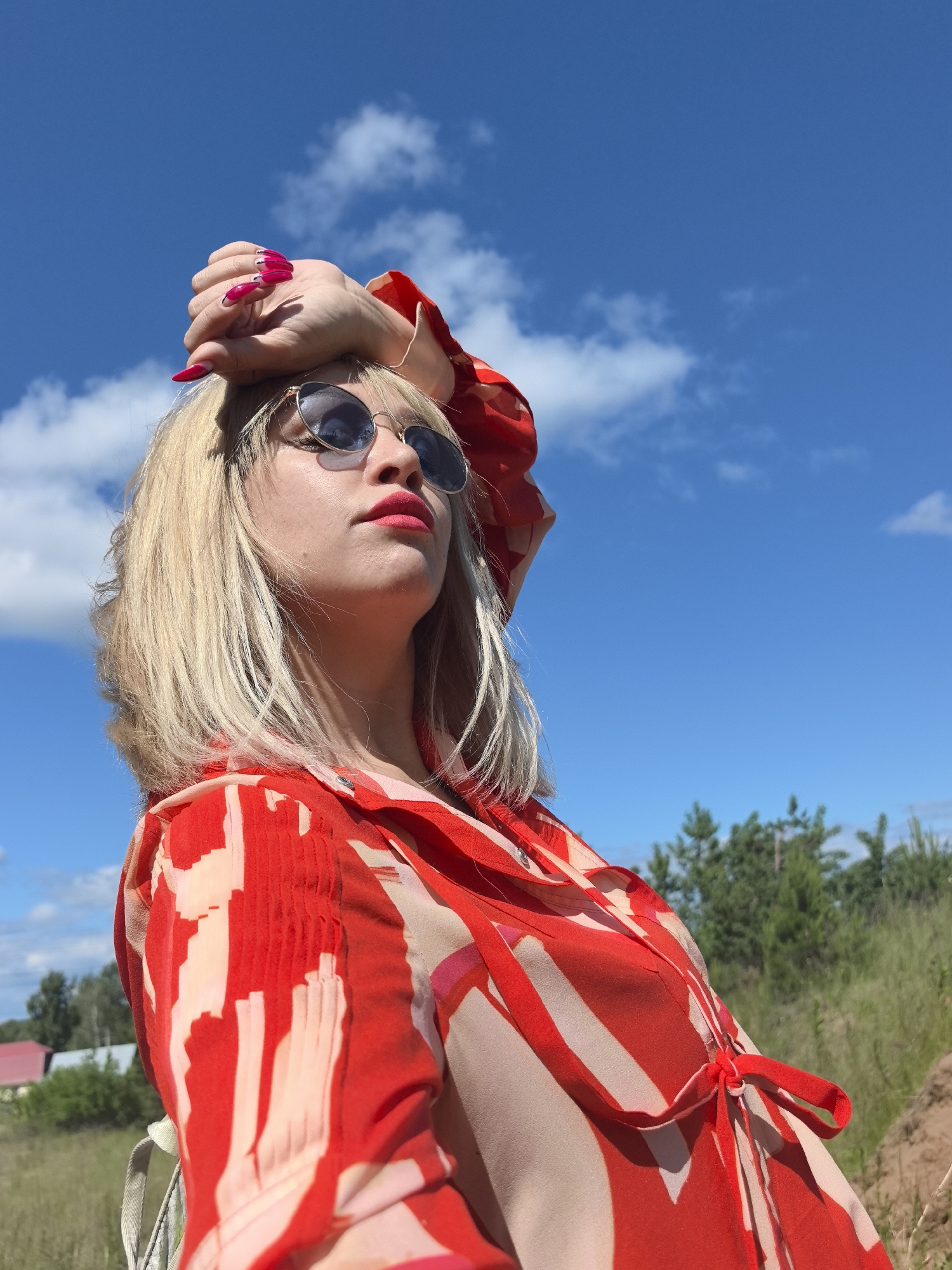 A low-angle, medium shot shows a woman with blonde hair and bright red lipstick, wearing sunglasses and a red and white patterned dress. She rests her right arm on her head, showcasing long, pointed red nails. The background is a bright blue sky with scattered clouds, and a grassy field with trees and a red-roofed building. The image was taken in the city of Noviy, Russia, likely outdoors in a rural or suburban area.