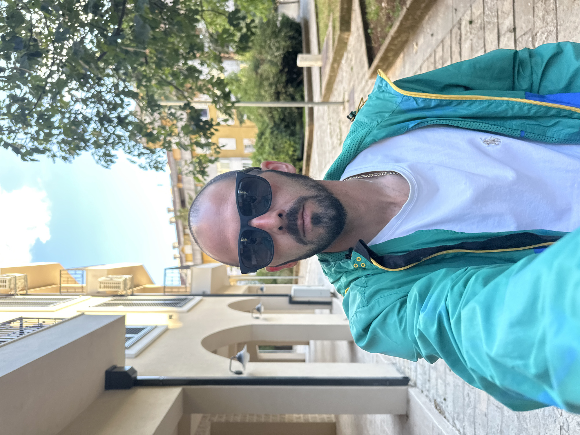 A man wearing sunglasses, a white T-shirt, and a green and blue jacket is standing in Budva, Montenegro. He is facing the camera. In the background, there are buildings with arches and trees. The sky is visible in the upper left corner of the image.