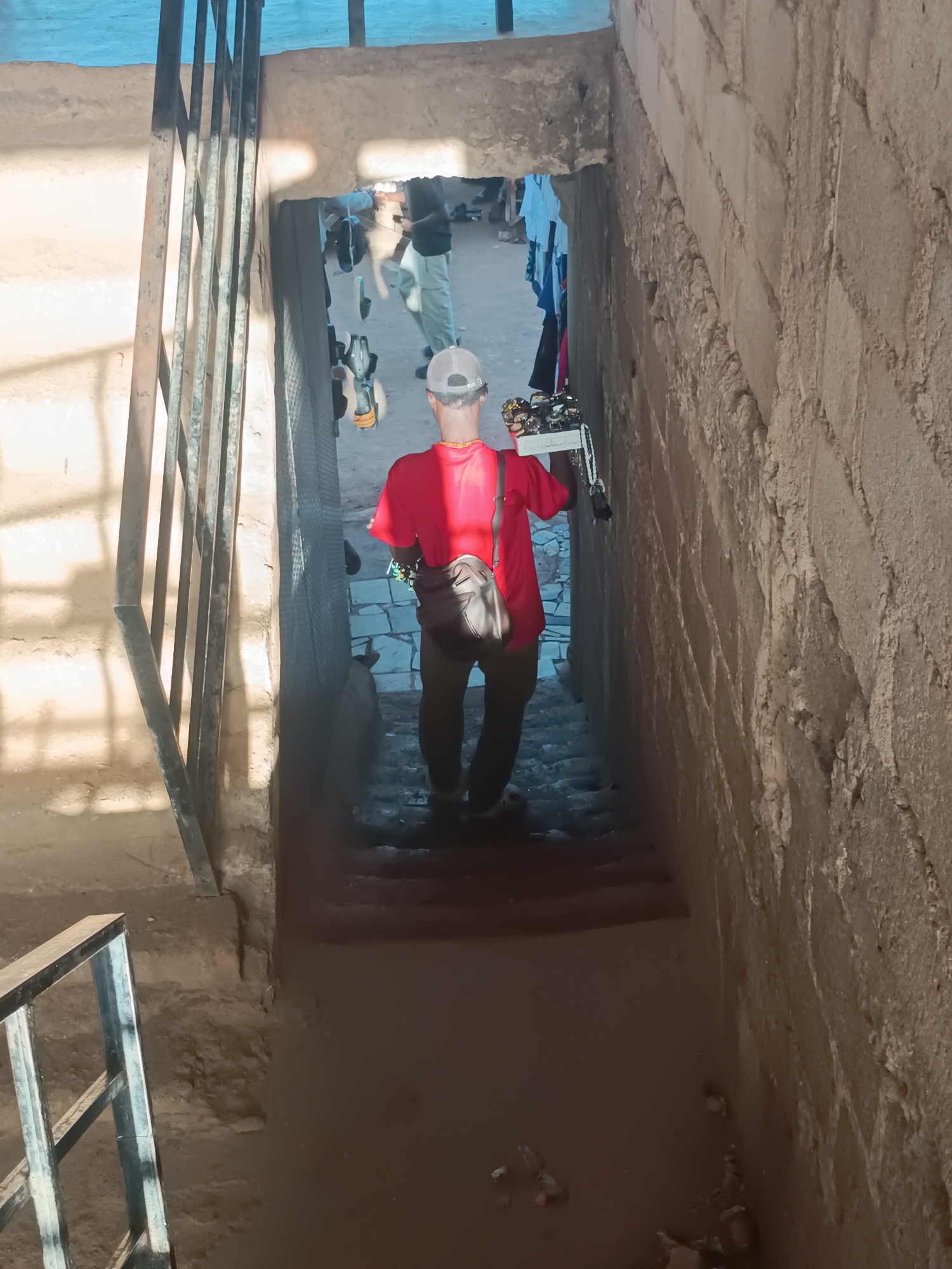 A man in a red shirt, tan trousers, and a white baseball cap is descending a flight of stone stairs with metal railings, exiting an opening with a view of an outdoor market scene. He carries a tray of goods, possibly sunglasses or jewelry, and has a shoulder bag. The walls of the stairwell are rough and textured. The location is Bukuru, Nigeria.
