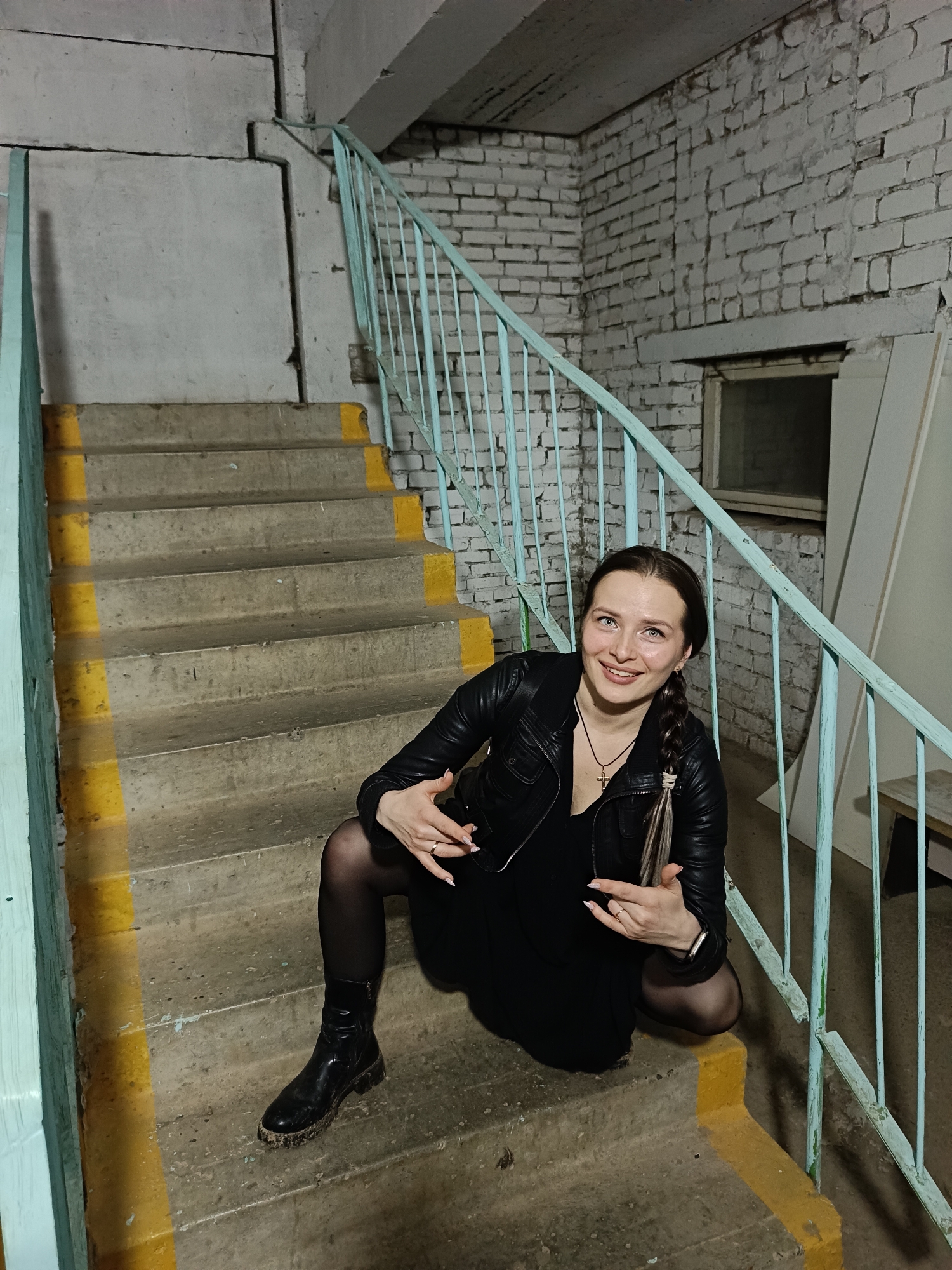 A woman poses for the camera in a stark, utilitarian stairwell in Chaikovsky, Russia. She is crouching on the third step from the bottom, looking up and to her right with a friendly smile. Her light brown hair is styled in a long braid with lighter streaks, extending over her left shoulder. She wears a black leather jacket over a black dress or top, black opaque tights, and chunky black boots that appear to have some dirt or debris on them. A silver cross necklace is visible around her neck, and she wears a ring on her left hand and a smartwatch on her left wrist. Her hands are gesturing outwards with open palms.
The setting is an indoor concrete and brick stairwell. The stairs are made of weathered concrete with bright yellow paint marking the edges of each step and the vertical risers. A light blue metal railing runs alongside the stairs on her right. The walls are a mix of painted white brickwork, revealing some wear and tear, and rough concrete slabs with visible seams and cracks. A small, obscured window is visible in the upper right portion of the background, and an open doorway or alcove is seen below it. The lighting is artificial and somewhat dim, casting shadows and highlighting the texture of the aged materials. The overall impression is that of a functional, possibly neglected, common area within an older building. There is no visible text in the image.