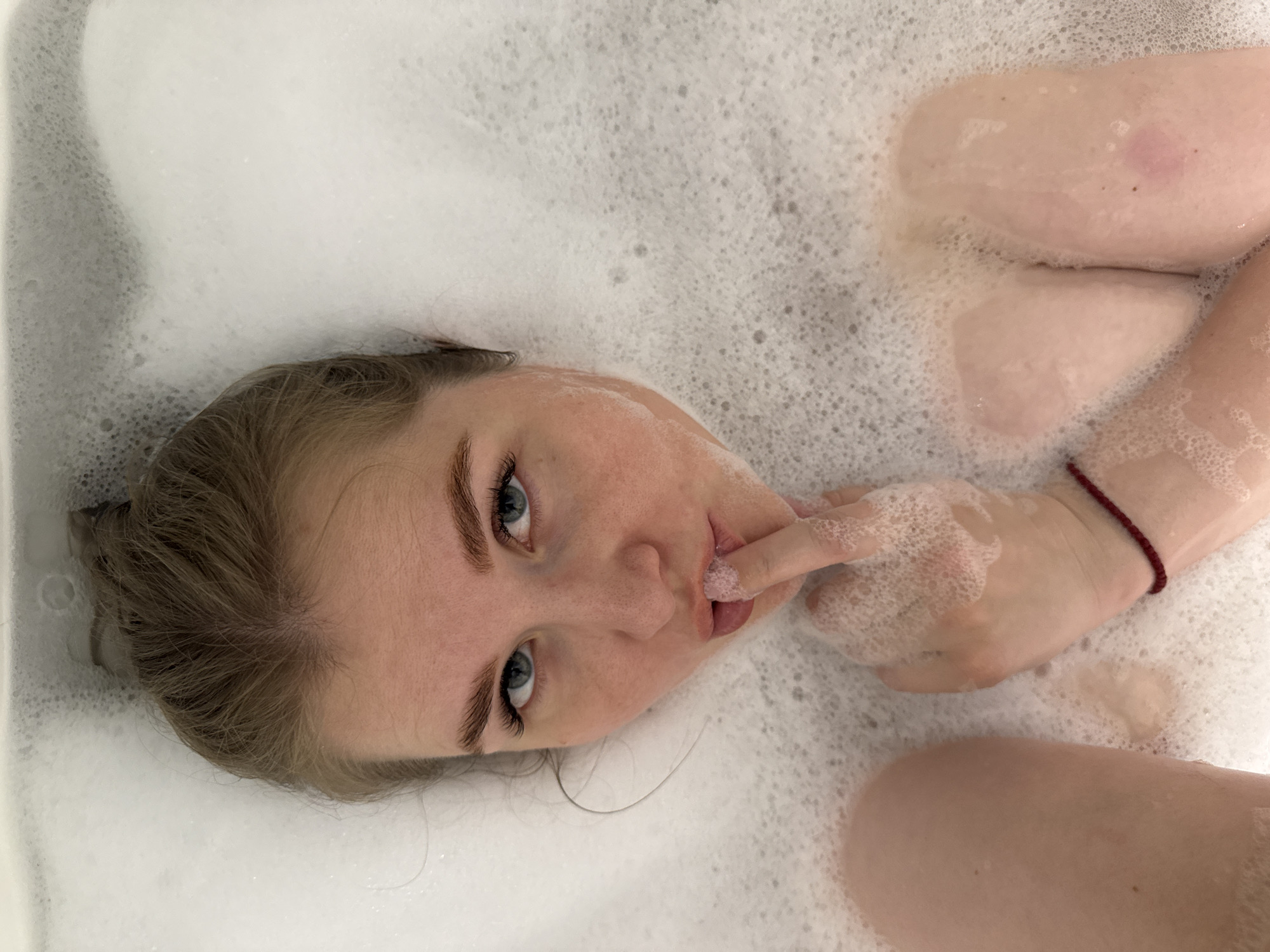 A fair-skinned person is in a bathtub filled with bubbles, the person is lying back in the tub and looking towards the camera. The person has light brown hair and blue eyes, and one finger is touching their lips. The person is wearing a thin, red bracelet on their right wrist. The bathtub appears to be white. There is no visible indication of the location being Ферма, Russia.