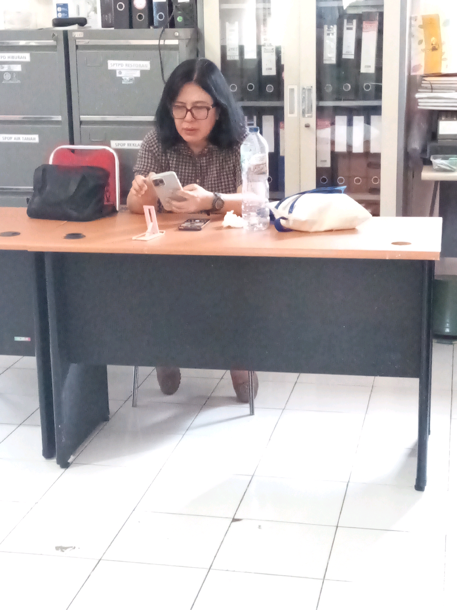 A woman sits at a wooden desk, holding a smartphone in her right hand. She is wearing glasses and a plaid shirt. On the desk are a bottle of water, a white pouch, and a small pink phone stand. The desk is in an office with filing cabinets and shelves filled with binders in the background. The floor is tiled.