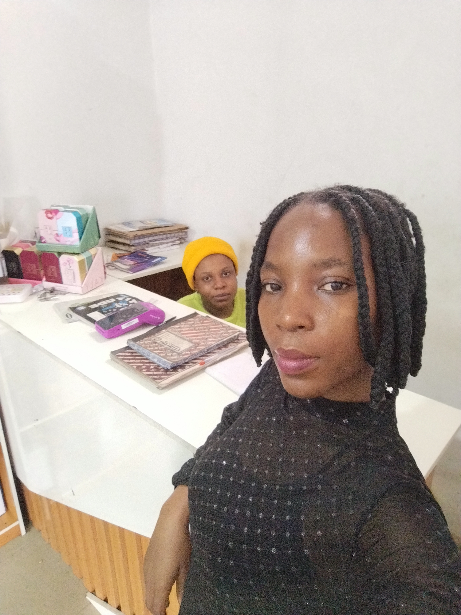 In Uselu, Nigeria, two women are visible behind a white counter. The woman in the foreground, facing the camera, has dark skin, braided hair, and is wearing a black, sequined shirt. She is positioned to the right of the frame, her head and upper body filling a significant portion of the image. The second woman, positioned behind and to the left of the first, has lighter skin and is wearing a yellow beanie and a green shirt. She is looking towards the camera.
On the counter, various items are arranged. To the left, there are stacks of colorful boxes, some with floral designs. Further to the right, multiple notebooks are visible, including one with a United States flag motif and another with an American flag design. A purple electronic device, possibly a card reader, rests on one of the notebooks. In the background, a stack of documents or books is placed on a shelf. The environment appears to be an indoor setting with white walls and a wooden front to the counter.