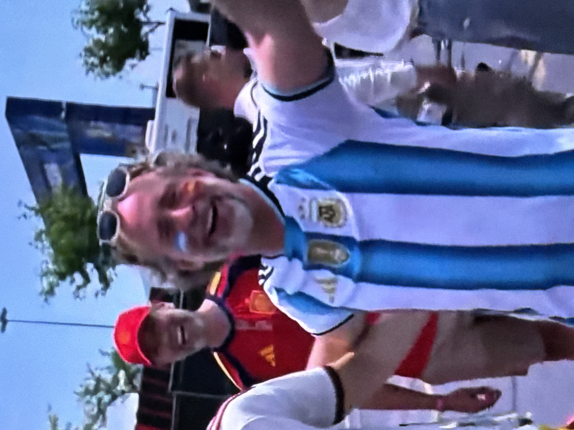 A man in an Argentina soccer jersey with blue and white stripes is pictured in the foreground, his face painted with blue and white stripes on his cheeks. He is smiling and wearing sunglasses on his head. To his left, a man in a red cap and a Spain soccer jersey is also smiling. In the background, there are trees, a blue structure, and other people. The scene appears to be outdoors in an urban or stadium environment.