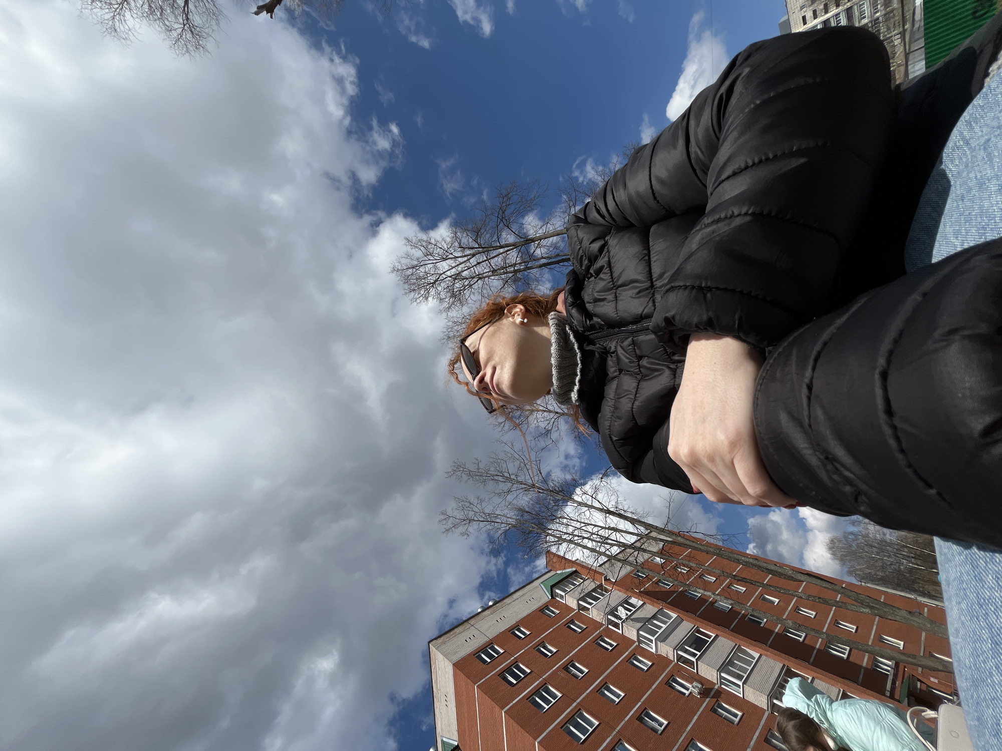 This image, taken in Chaikovsky, Russia, is an outdoor shot captured from a low, upward-looking angle. The main subject is a person with reddish-brown, curly hair, visible in profile on the right side of the frame. They are wearing black sunglasses, a black puffer jacket, and a grey collar or scarf, and appear to be looking upwards or into the distance. A portion of their light blue jeans is also visible.
In the bottom right corner, the back and head of a second person, wearing a light blue jacket or hoodie, are partially visible, looking downwards.
The background is dominated by a dynamic sky, featuring patches of clear blue with scattered white cumulus clouds, alongside areas of more uniform grey clouds. Bare tree branches, suggesting late winter or early spring, extend across the sky. A large, multi-story apartment building, constructed from red brick with numerous white-framed windows and balconies, occupies the lower right and center of the frame, its architecture consistent with urban settings in Chaikovsky. The scene is brightly lit, indicating daytime. No visible text is present.