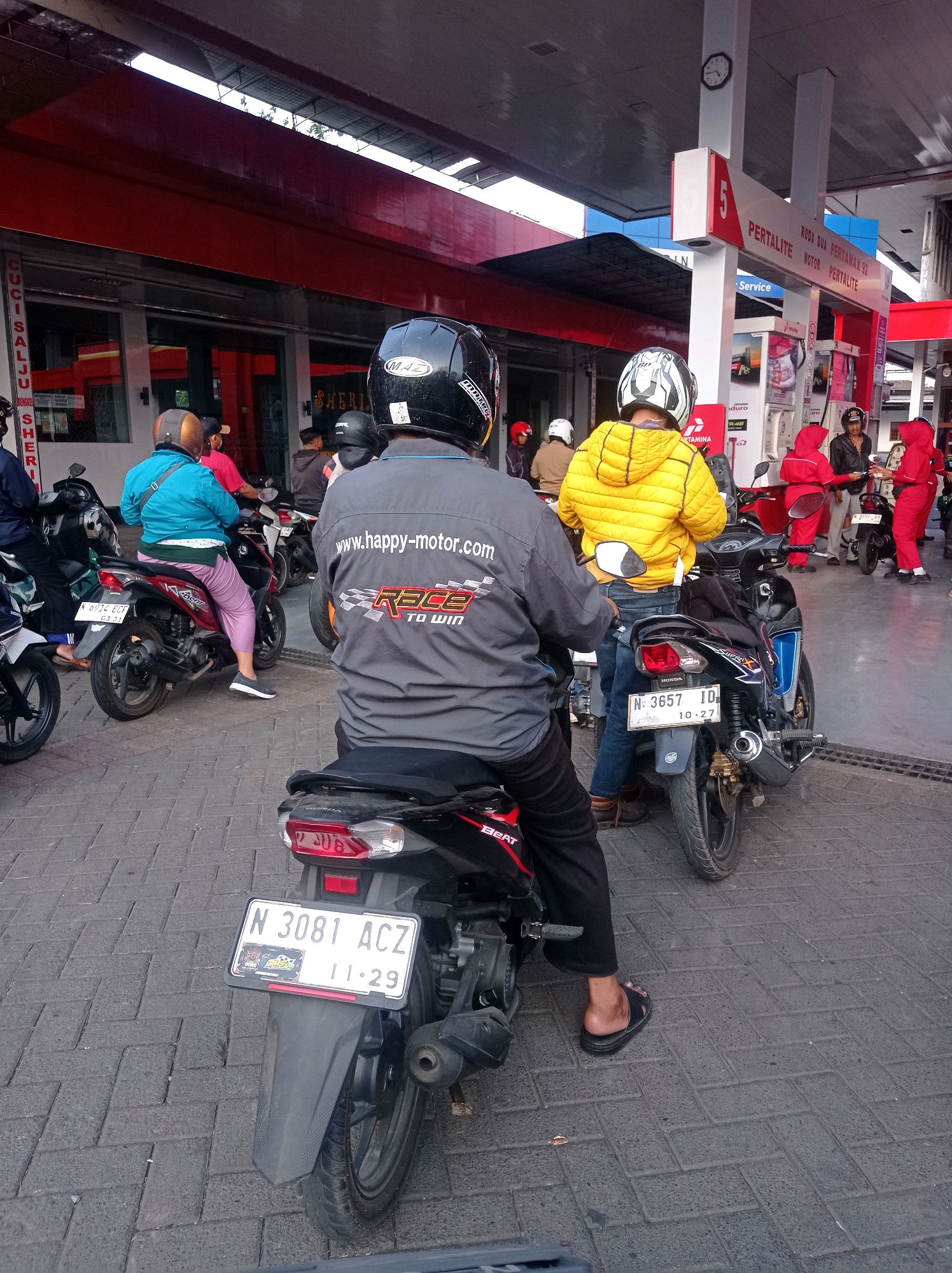 The image depicts a queue of motorcyclists at a Pertamina gas station in Bululawang, Indonesia. The foreground shows the rear of a motorcycle with the license plate "N 3081 ACZ" and a date stamp of "11-29". A person wearing black pants and black sandals is seated on this motorcycle, facing away from the camera. Behind them, another person in a yellow jacket and blue jeans is on a motorcycle. Further in the background, individuals in red uniforms, presumably gas station attendants, are interacting with customers. The gas station features red and white signage, including "PERTALITE" and "RODA DUA". The ground is paved with grey interlocking bricks. The environment suggests a typical urban setting with commercial establishments.