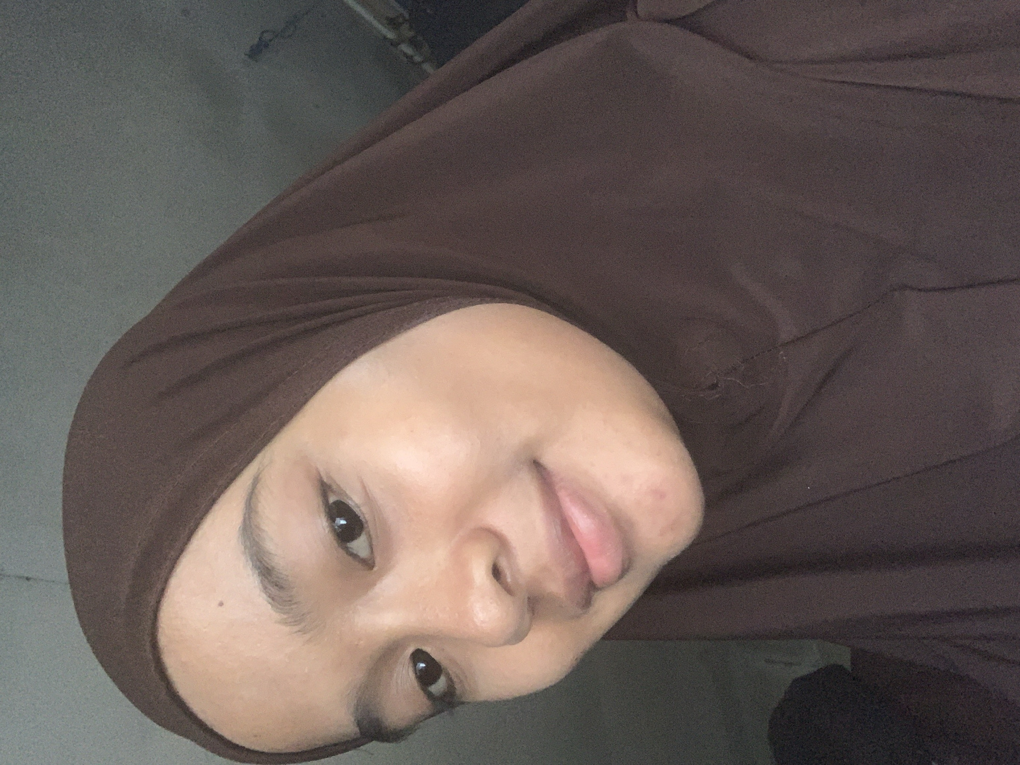 A close-up, chest-up, indoor selfie shows a young Indonesian woman from Sungai Meriam, Indonesia. She is wearing a brown hijab and has a light complexion. Her eyes are dark brown and her lips are light pink. Her head is tilted slightly to the left and she is smiling. The background is out of focus and appears to be a wall. The lighting is dim.