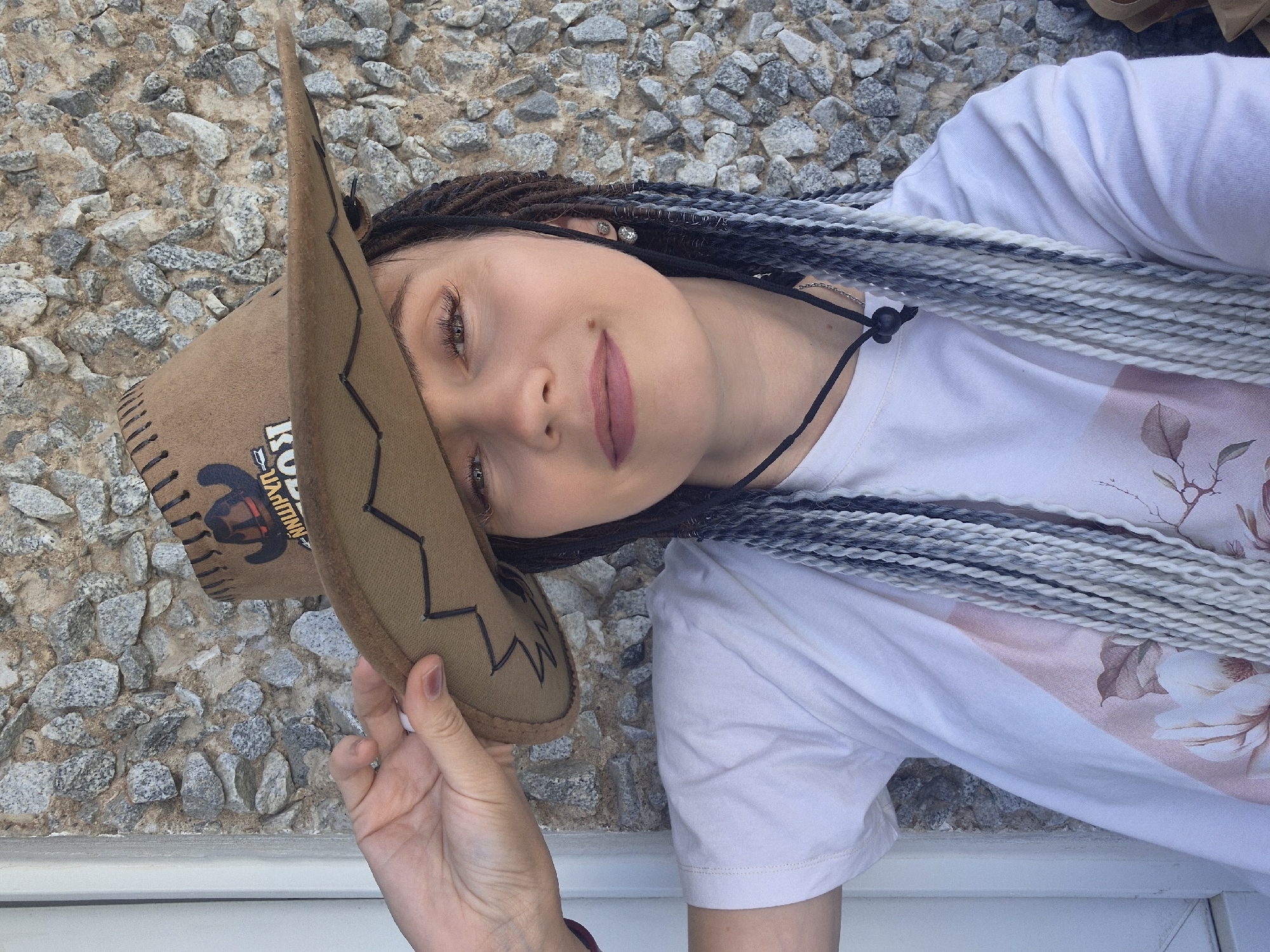 A young woman with braided hair, varying in color from dark brown to white, is posing for a selfie outdoors. She is wearing a white t-shirt with a floral print. On her head, she has a brown cowboy hat which she is holding with her right hand. The background is a stone wall. The scene appears to be in Chaikovsky, Russia.