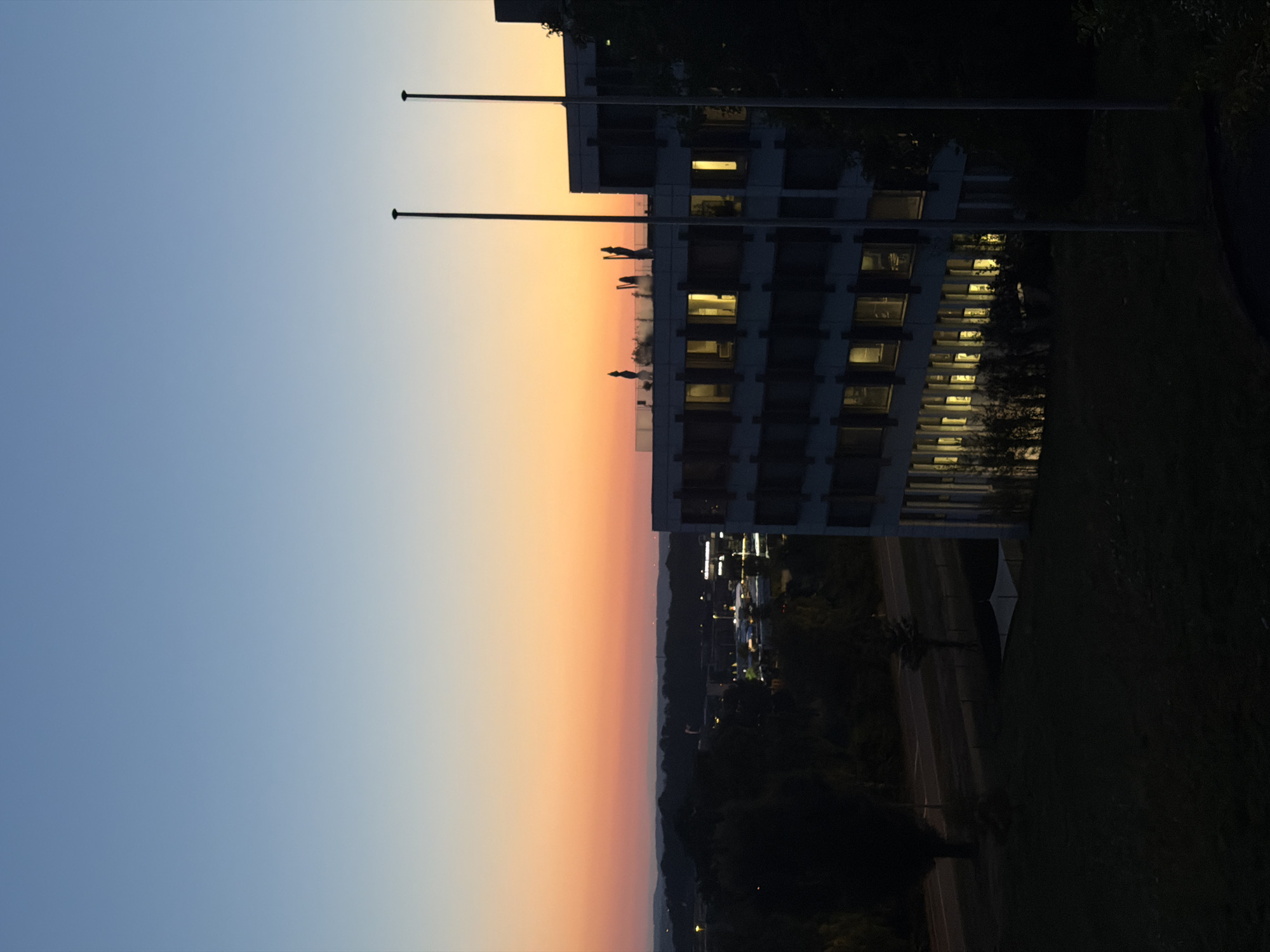 The image depicts a twilight scene in Leudelange, Luxembourg, showing a modern, multi-story building illuminated from within. The sky transitions from a clear, pale blue at the top to warm orange and pink hues near the horizon, indicating sunset or sunrise. In the foreground, a dark, grassy area leads towards a road and more buildings in the middle ground. The architecture of the main building features a repetitive pattern of windows, some of which are brightly lit, suggesting occupancy or internal lighting. Two tall flagpoles stand prominently in front of the building. The overall atmosphere is calm and serene, with the fading light casting a gentle glow over the urban landscape.