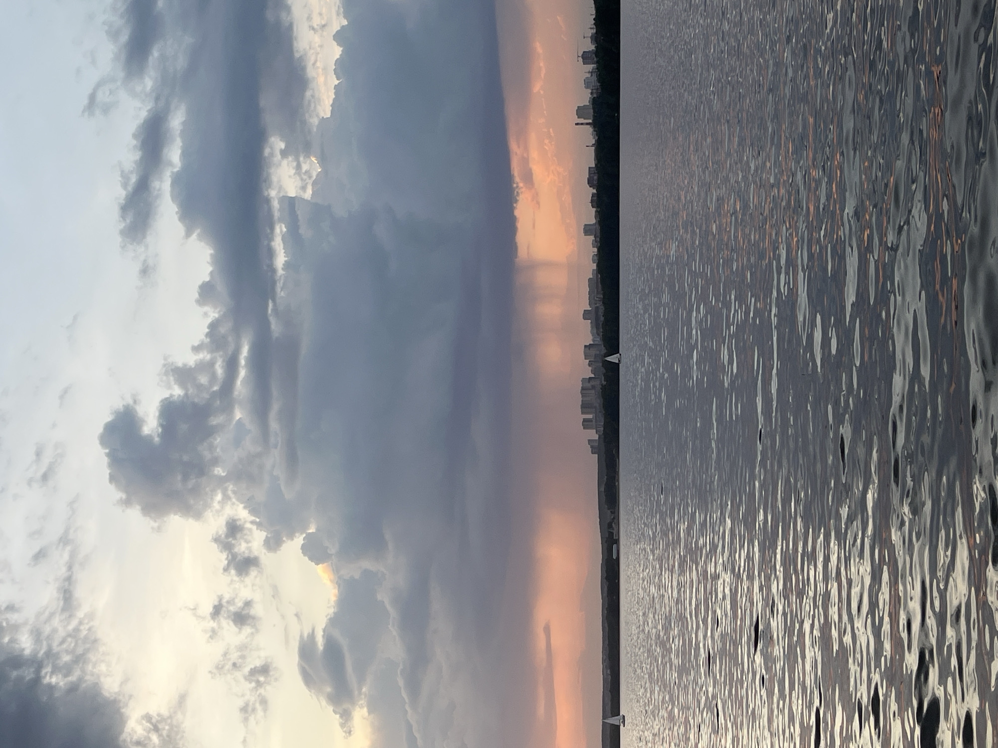 The image shows a body of water, likely the Iset River in Yekaterinburg, Russia, with cityscape silhouettes visible on the horizon. The sky features a mix of dark, stormy clouds and areas with a peachy, sunset-like glow. The water's surface reflects the colors of the sky. A small sailboat is visible on the water. The image is oriented in a landscape format, though it appears to be taken in portrait mode and then rotated.