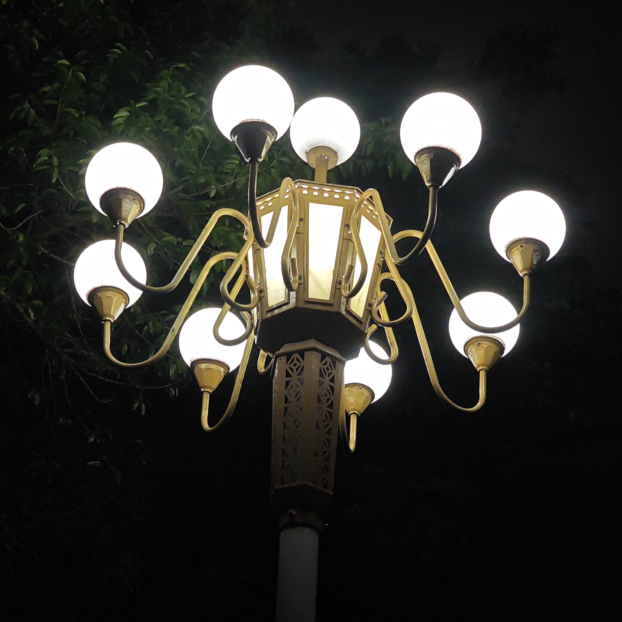 A streetlight with a central lantern and eight spherical lamps is situated against a backdrop of trees at night. The streetlight's body features decorative lattice panels and is mounted on a plain cylindrical pole. The lamps are illuminated, and the scene is framed by the darkness of the night sky. Based on the image, the location in the city of Yuzhong District, China cannot be determined.