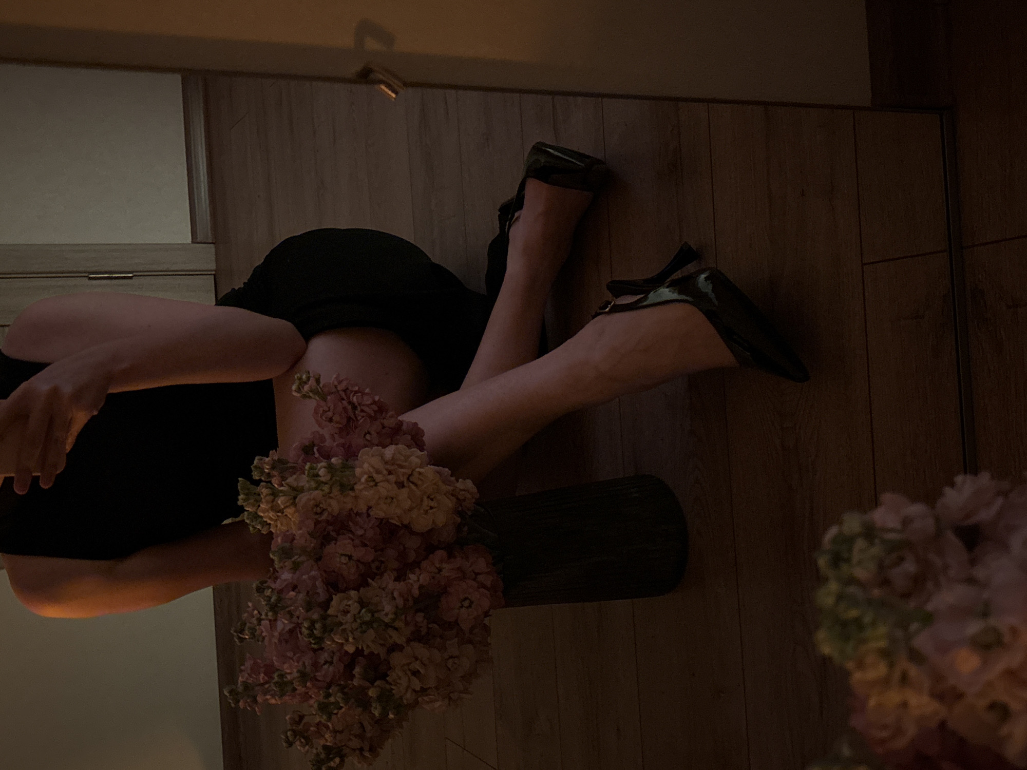 The image captures a person's legs and arms, dressed in a black outfit and black pointed-toe heels, as they lie on a wooden floor. A bouquet of light pink and cream flowers sits in a dark vase next to the person's legs. The scene appears to be indoors, with a reflection of furniture and a wall visible in the background. The lighting is dim and warm, suggesting an evening or indoor setting. The overall mood is calm and possibly intimate, with the focus on the details of the person's attire and the floral arrangement. No text is visible in the image.