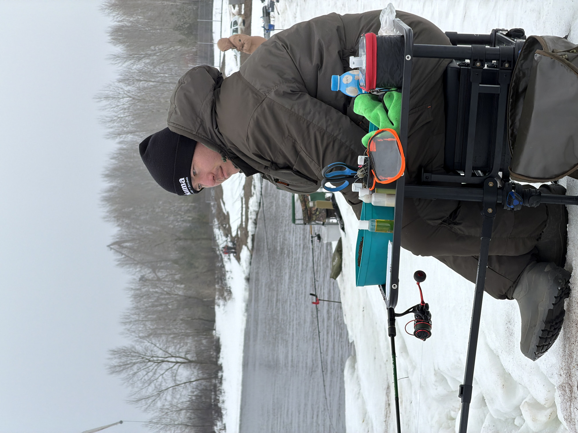 The image depicts a man fishing in a snowy, outdoor environment. The man is dressed in a dark green parka with the hood up, a black Puma knit cap, and grey boots. He is leaning over a fishing platform which holds a variety of items including a water bottle, green gloves, scissors, containers of bait or attractant, and a teal blue box containing dark brown bait. A fishing rod with a red and black reel is set up in the snow. The background shows a body of water, likely a river or lake, with snow-covered banks and bare trees along the opposite shore. There is also what seems to be a building in the background. The scene is set in a winter landscape, with the ground covered in snow and the sky overcast. The image is consistent with the city of Chaikovsky, Russia, given the climate and general environment depicted.