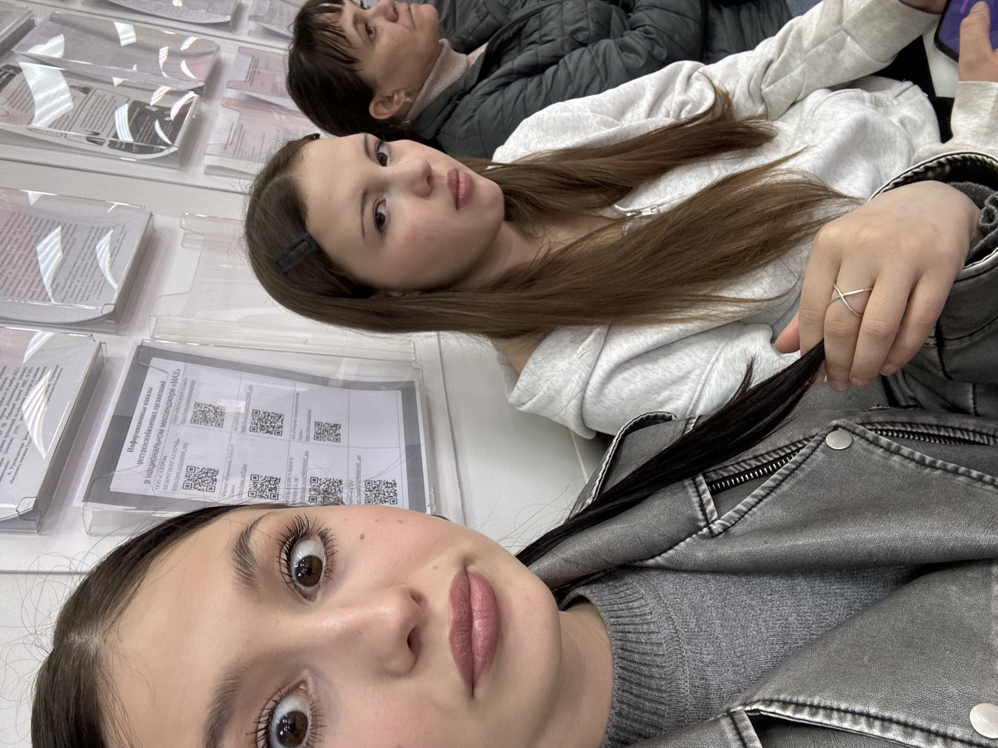 The image captures three females in an indoor setting, likely a public service center or waiting room, in Chaikovsky, Russia. The shot is taken as a selfie, with the closest subject, a young woman with dark hair, prominent in the foreground. She has wide brown eyes, defined eyelashes, and pink/mauve lipstick, wearing a gray knitted sweater and a gray faux leather jacket. Her expression is attentive towards the camera. Behind her, a second young woman with long light-brown hair, wearing a light gray hooded sweatshirt and a silver ring, looks towards the camera with a slight pout. The third person, an adult woman with short dark hair and a dark quilted jacket, is visible in the background, looking upwards and away from the camera with a neutral expression.
On the white surface adjacent to them, several documents are displayed in clear plastic holders. These documents feature Russian text and multiple QR codes, suggesting information related to services. Visible text includes "Информационные каналы ресурсоснабжающих организаций в национальном мессенджере «МАХ»" (Information channels of resource-supplying organizations in the national messenger "MAX"), along with mentions of "ООО «ГАЗПРОМ МЕЖРЕГИОНГАЗ ПЕРМЬ»" (Gazprom Mezhregiongaz Perm LLC) and "ПАО «ЛЕРМОНЕРГОСЬО»". The scene conveys a casual, candid moment as two of the subjects pose for the photograph.