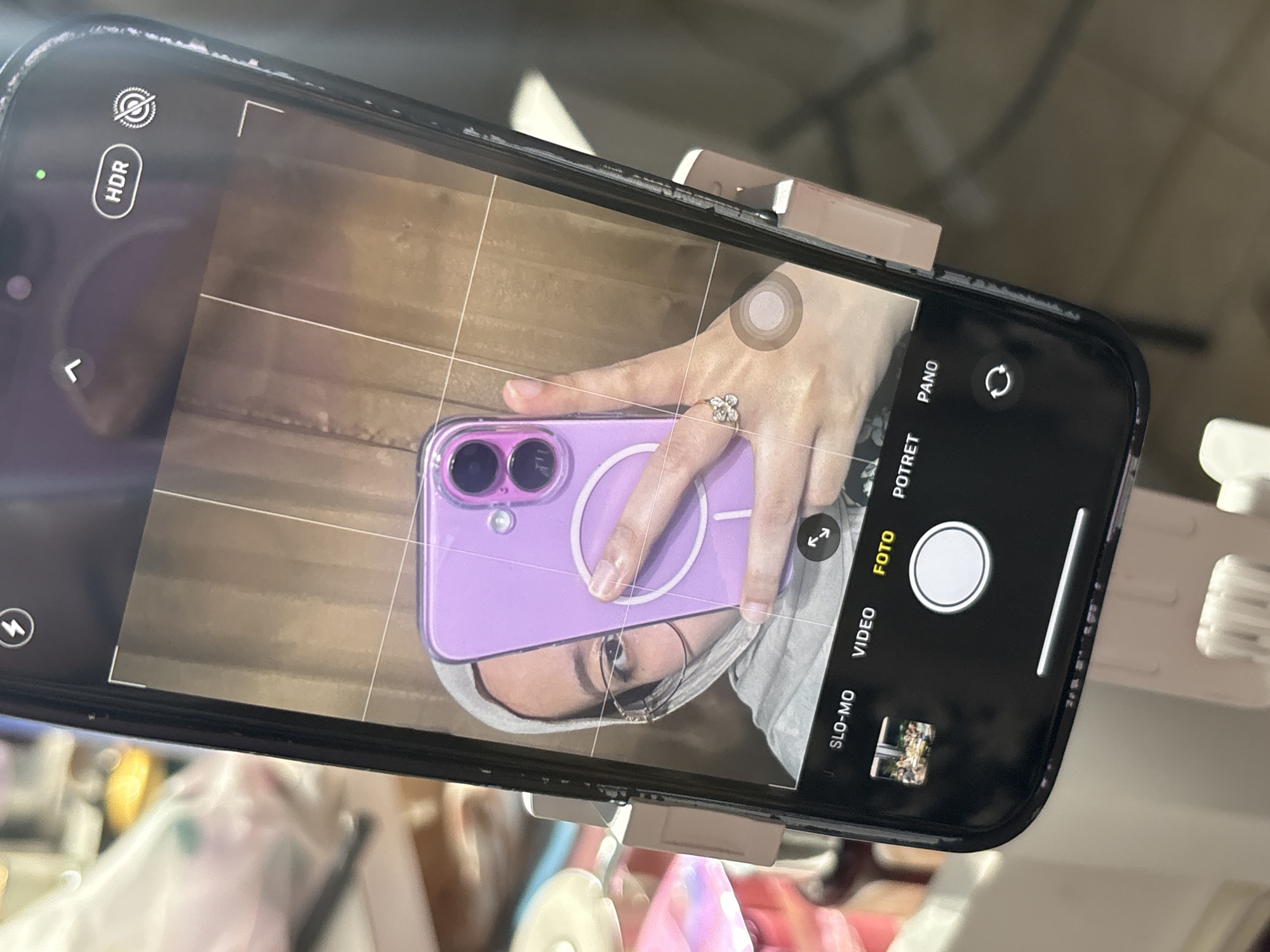 The image shows a black smartphone held by a clip, with its camera app open, aimed at capturing a person (likely female, identifiable by the eye, eyebrow, skin tone and wearing glasses) holding a purple smartphone. The background visible in the camera's viewfinder shows a wooden surface and what appear to be fingers with a ring. The phone is in a cluttered location with other unidentifiable objects in the foreground. No specific location in the city of Gadingrejo, Indonesia can be determined from the image.