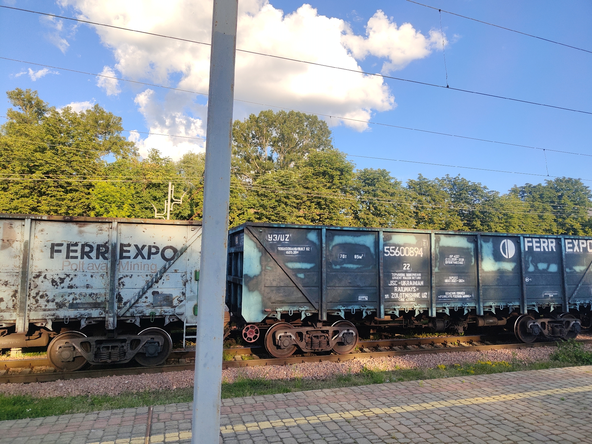 A high-angle, wide shot captures a sunny day with blue skies and scattered white clouds above a train parked on tracks. The train consists of two freight cars, the first appearing weathered and the second a darker teal color with various markings. The first freight car displays the words "FERRI EXPO Poltava Mining" in large, dark letters. The second freight car features numerical designations such as "55600894" and "22," along with text in Cyrillic script. Train tracks run horizontally across the lower portion of the frame, bordered by gravel and grass. A paved walkway with a yellow and gray checkered pattern is visible in the foreground. Dense green trees line the background, suggesting a park or natural area. A slender, light gray vertical pole is positioned in the center of the image, partially obscuring the train. The scene appears to be set at a train station or siding within Kyiv, Ukraine, based on the presence of the train and general urban/suburban environment.