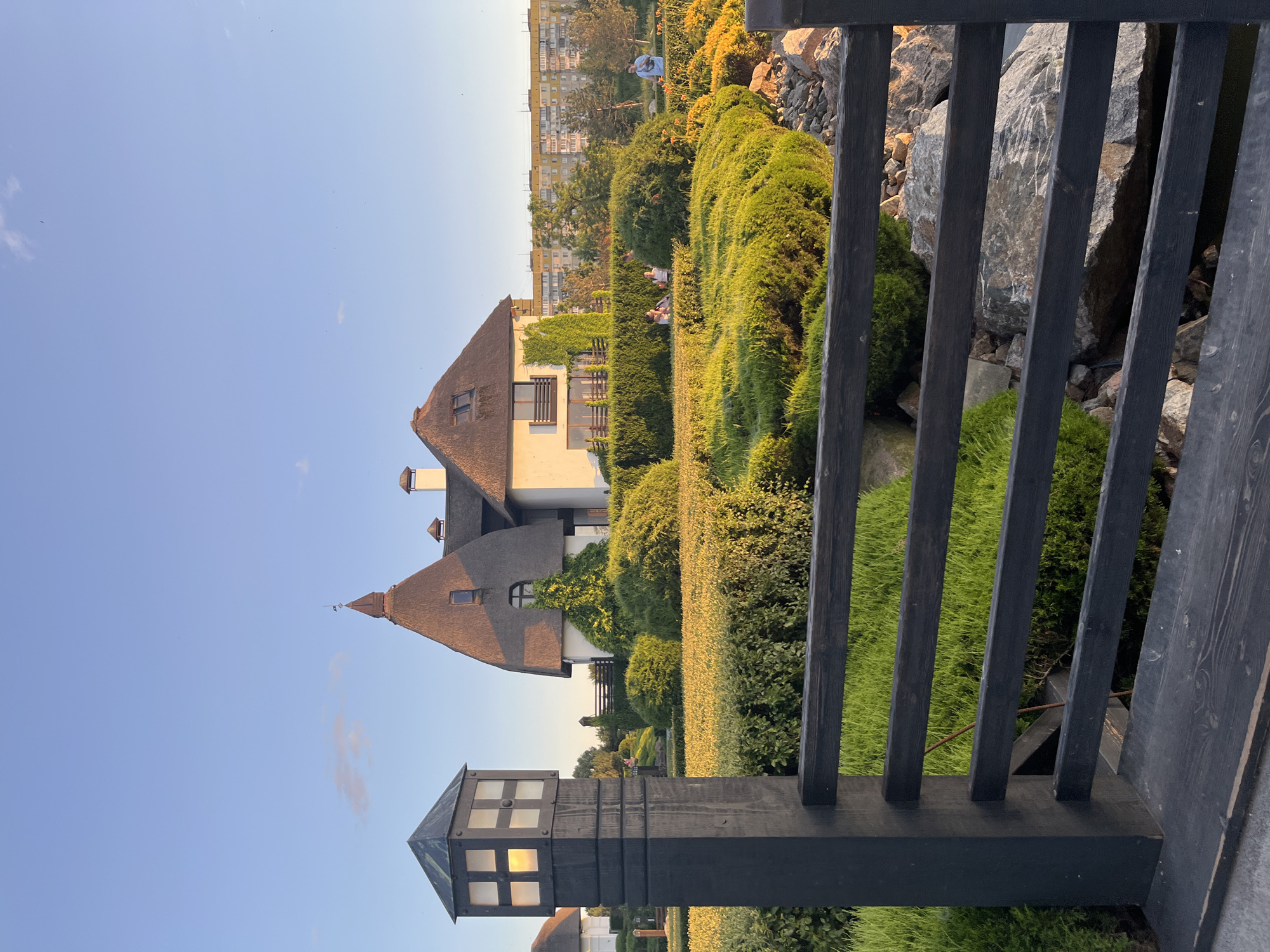 In the city of Dnipro, Ukraine, at the Amur district, a photograph captures a residential building with a thatched roof and a distinct pointed turret, bathed in the warm light of a sunset. The building is surrounded by meticulously landscaped gardens, featuring neatly trimmed hedges and vibrant green grass. A dark wooden fence with horizontal planks separates the foreground from the garden. A black wooden post with a lantern-like light fixture stands to the left of the fence. In the background, a multi-story apartment building is visible. Several individuals are present in the garden, some seated and others standing, appearing to enjoy the evening. The sky is clear blue, with a few wispy clouds, indicating fair weather.