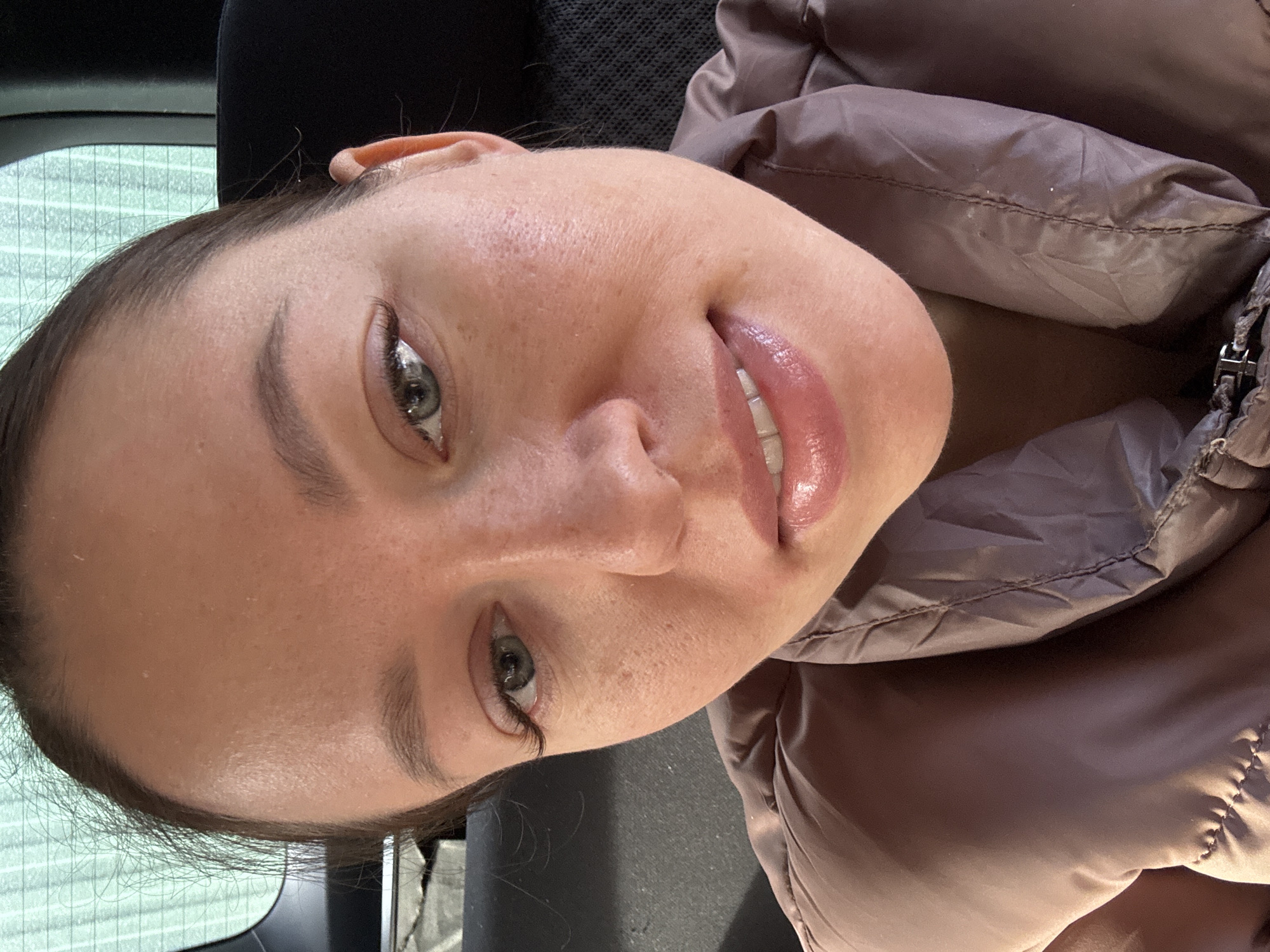 The image depicts a close-up, head-and-shoulders portrait of an adult female located within a vehicle in the city of Ферма, Russia. She has a light complexion, green-grey eyes, and dark brown hair pulled back, with some loose strands visible around the forehead. Her lips are glossy, and she displays a subtle, closed-mouth smile while gazing directly at the camera. She is dressed in a light brown, quilted puffer jacket. The background shows the interior of a car, specifically the rear window with vertical defroster lines visible on the upper left, and a dark, fabric-covered headrest or seat portion to the right.