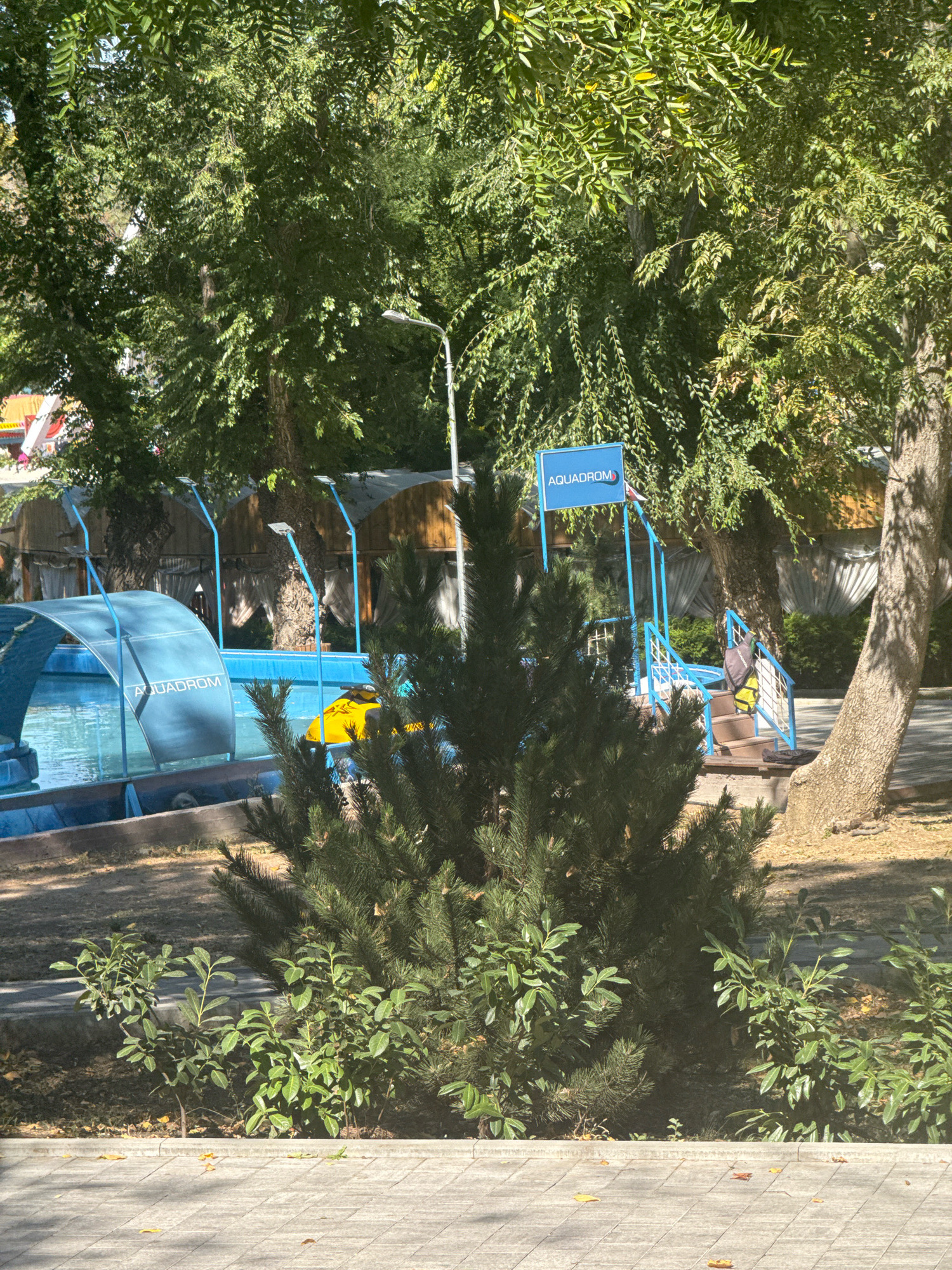 A sunny outdoor scene at an aquapark in Anapa, Russia, features a blue and white slide structure next to a pool. A bright yellow inflatable object is partially submerged in the pool. A sign that reads "AQUADROM" is attached to the blue structure. Various trees with lush green foliage provide shade. A concrete pathway runs in the foreground, with scattered fallen leaves. The overall impression is of a recreational area on a clear day.