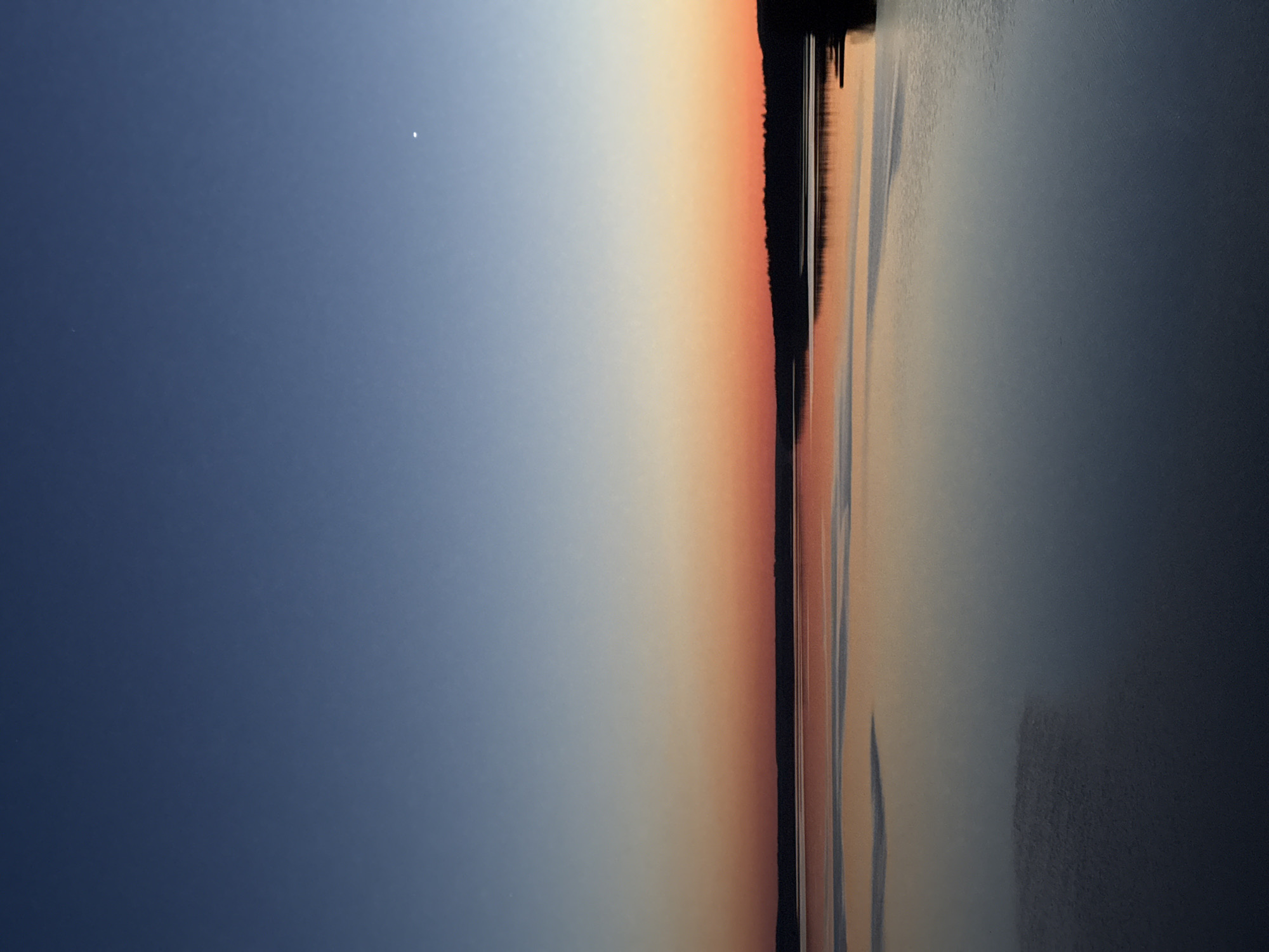 The image captures a landscape at sunset or sunrise, likely near a body of water. The sky transitions from dark blue on the left to a vibrant orange-red near the horizon. A dark, silhouetted landmass or trees line the horizon, possibly representing a shoreline. The water reflects the colors of the sky, creating a mirrored effect. The exact location in Votkinsk, Russia cannot be determined from the image alone. There are no people, discernible objects, actions, or interactions visible in the image beyond the natural elements of the sky, land, and water.