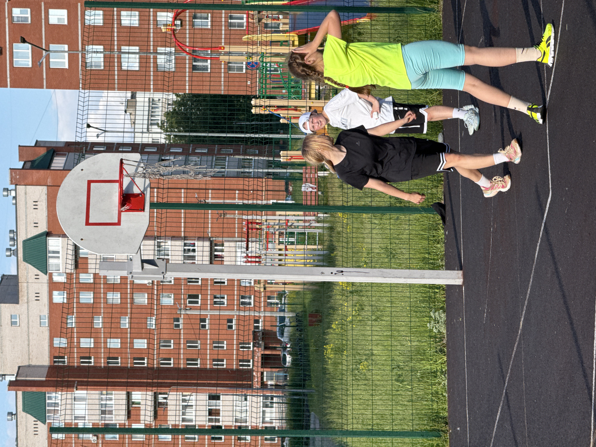 In Chaikovsky, Russia, on a sunny day, three individuals are present on an outdoor basketball court. Two young women and one young man, dressed in athletic wear, are engaged in an activity on the black asphalt court, marked with white lines. One woman, wearing a black t-shirt and shorts, is in motion. Another woman, in a bright yellow t-shirt and light blue shorts, stands to the right, with her hand raised to her head. The young man, in a white t-shirt and shorts, stands between them, holding a small white object. A basketball hoop with a net is visible in the background, along with a green metal fence. Beyond the fence, there is a grassy area with yellow flowers and several multi-story brick apartment buildings, characteristic of urban Chaikovsky. The scene suggests a casual game or practice session.