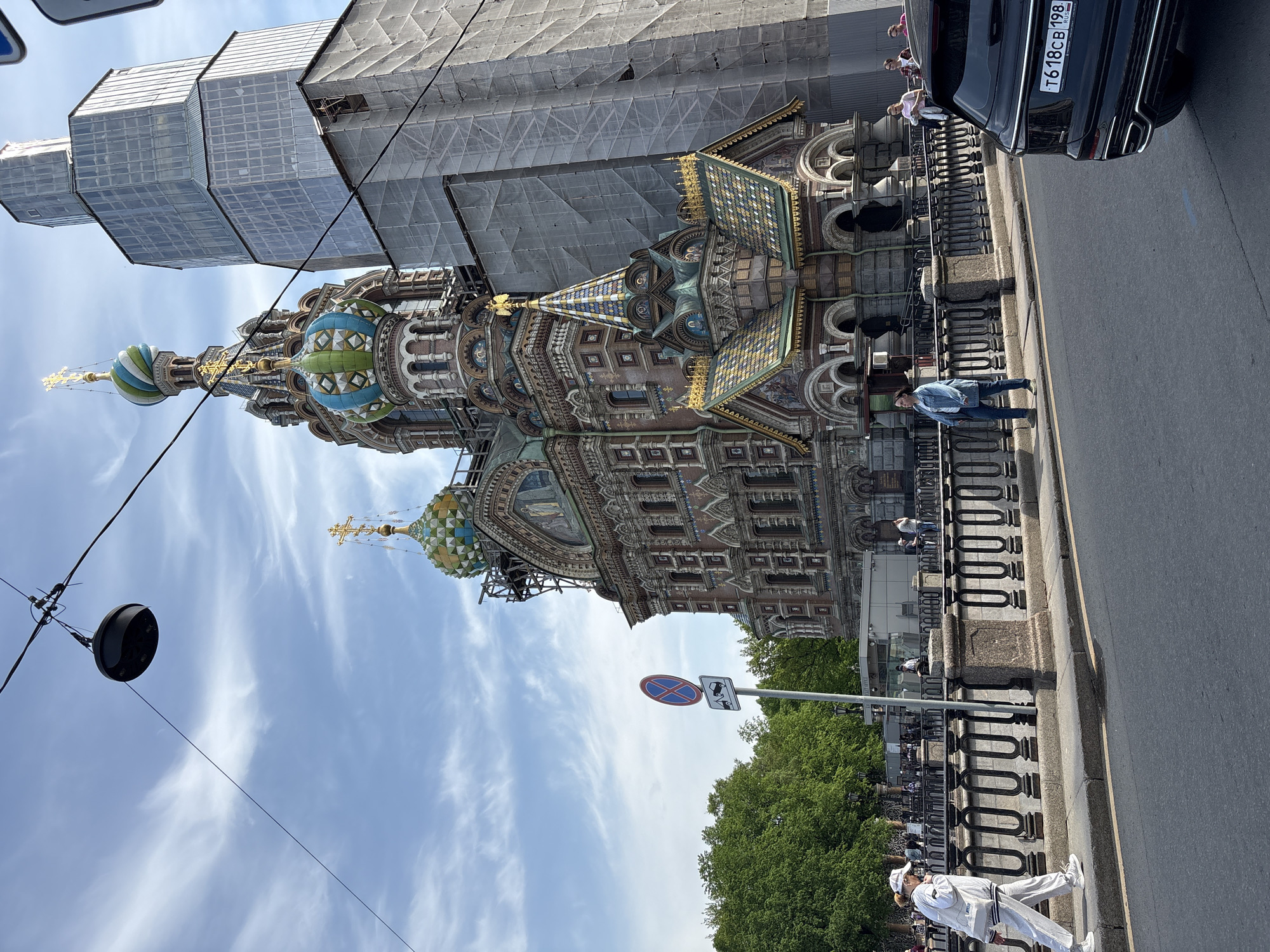 The Church of the Savior on Spilled Blood in St. Petersburg, Russia, stands prominently along a road with a black vehicle parked to its right. Scaffolding partially covers the building. Several people are present, including a person walking along the sidewalk dressed in white and others standing by the fence. A street light fixture hangs from a pole in the foreground. The sky above features scattered clouds. A road sign is visible along the sidewalk.