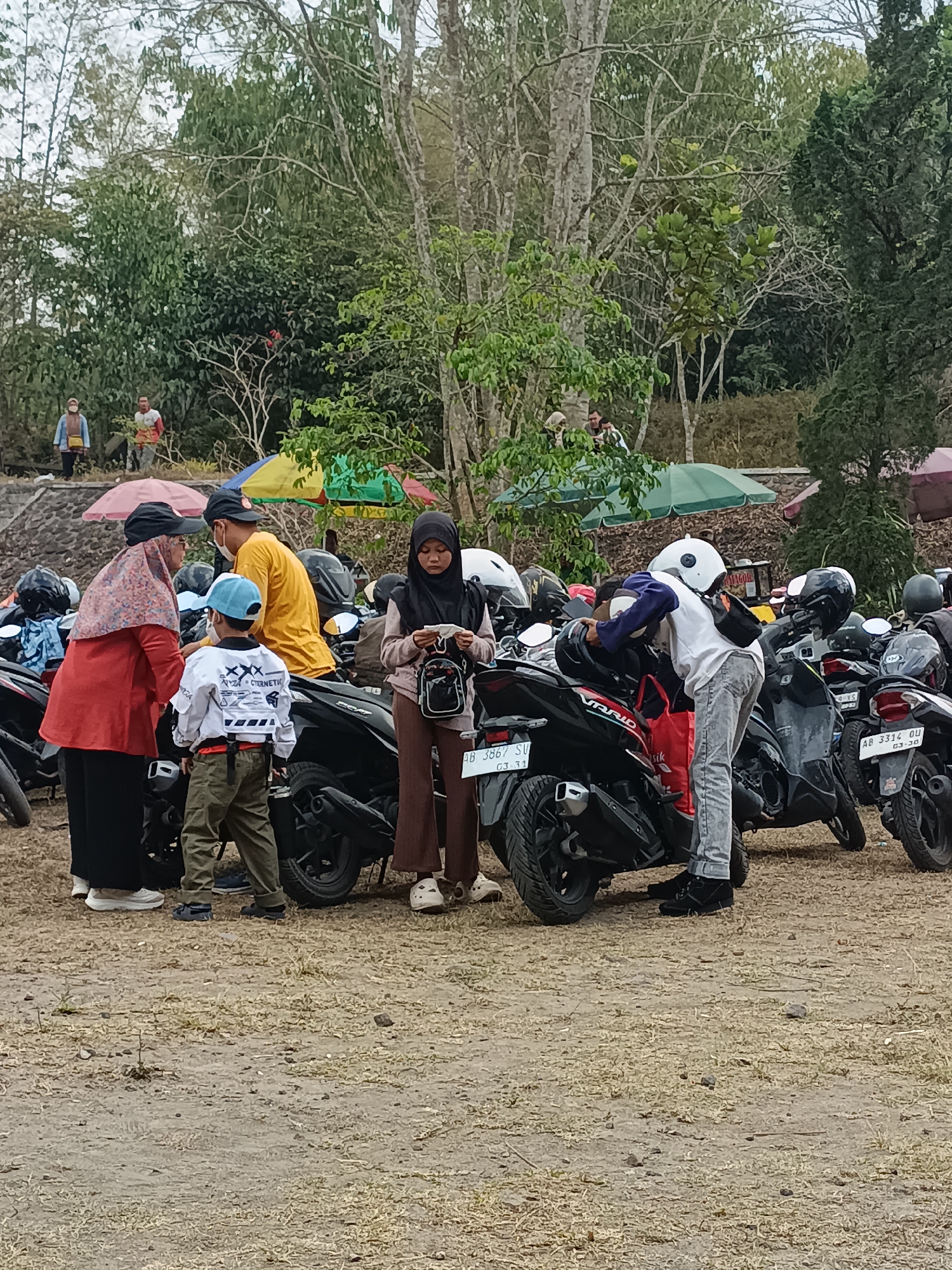 A group of people are gathered around parked motorcycles in what appears to be an outdoor parking area. The setting includes dry, patchy grass and a dirt ground. In the background, there are trees with lush green foliage and some taller, bare trees, indicating an outdoor, possibly rural or park-like environment. There are also colorful umbrellas visible, suggesting a possible recreational area or event.
Specifically within Hargobinangin, Yogyakarta, Indonesia, this scene could be a parking lot at a tourist attraction such as the Kaliurang area of Mount Merapi or another natural park.
The individuals present include adults and a child. One woman in the foreground wears a red top and a patterned headscarf, interacting with a young boy in a white shirt and green pants. A man in a yellow shirt stands nearby. Another woman in a black headscarf is seen looking at her phone. A person in a white t-shirt and grey pants is seen leaning over a motorcycle. Several motorcycles are parked closely together. The people are dressed casually. The interaction between the people appears casual, with some engaged in conversation and others attending to their belongings or motorcycles.
