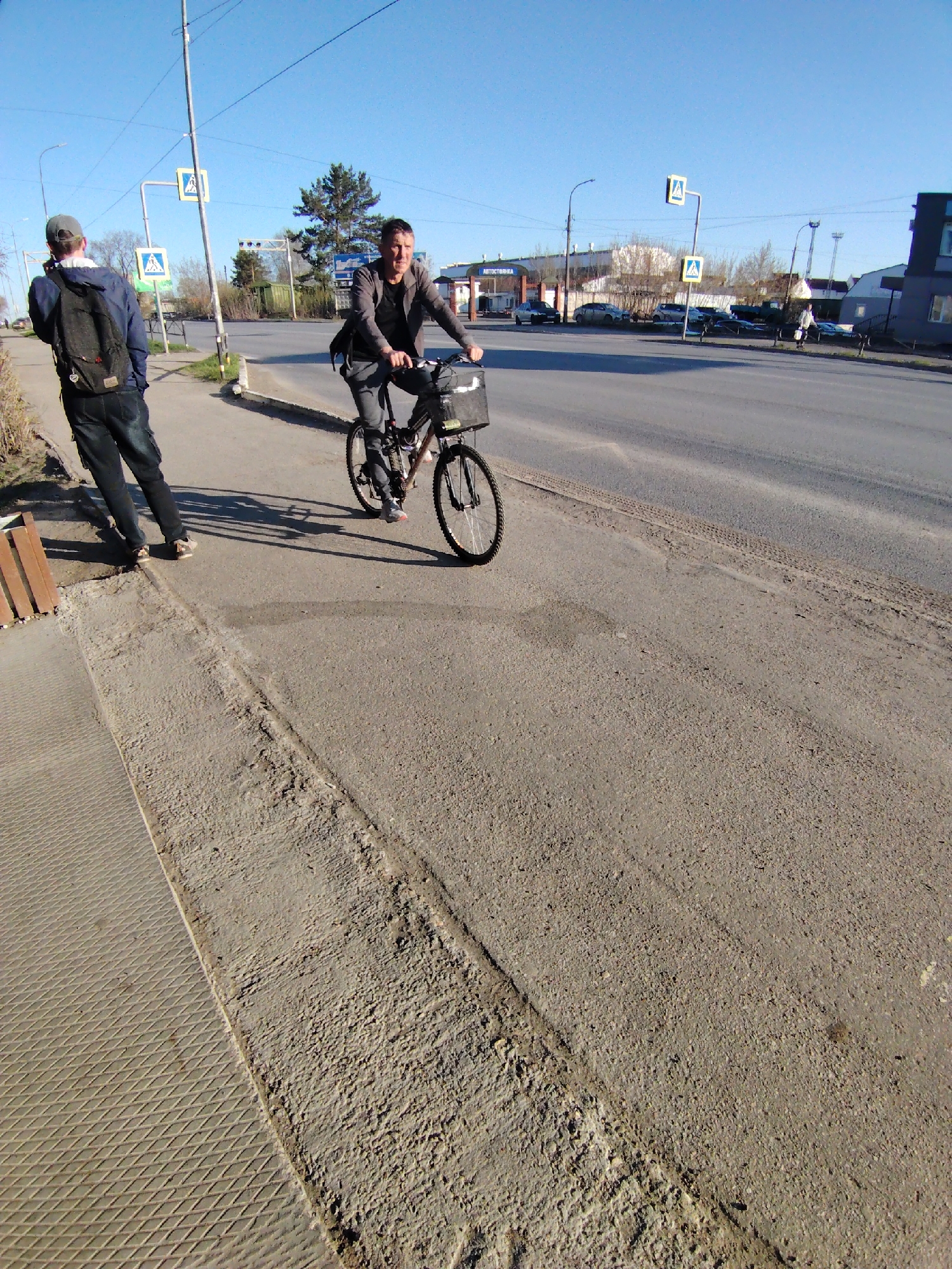 A medium, sunny, wide outdoor shot reveals a man riding a bicycle along a street in what appears to be the city of Chaikovsky, Russia. Another man stands on the sidewalk with a backpack. The bicyclist wears a dark jacket and trousers. His bicycle has a basket on the front. The pedestrian is dressed in a blue jacket, dark trousers, and a baseball cap. In the background is a street with parked cars and buildings. Pedestrian crossing signs are visible. The road shows signs of wear and tear. The overall impression is that of an everyday scene in a Russian city.