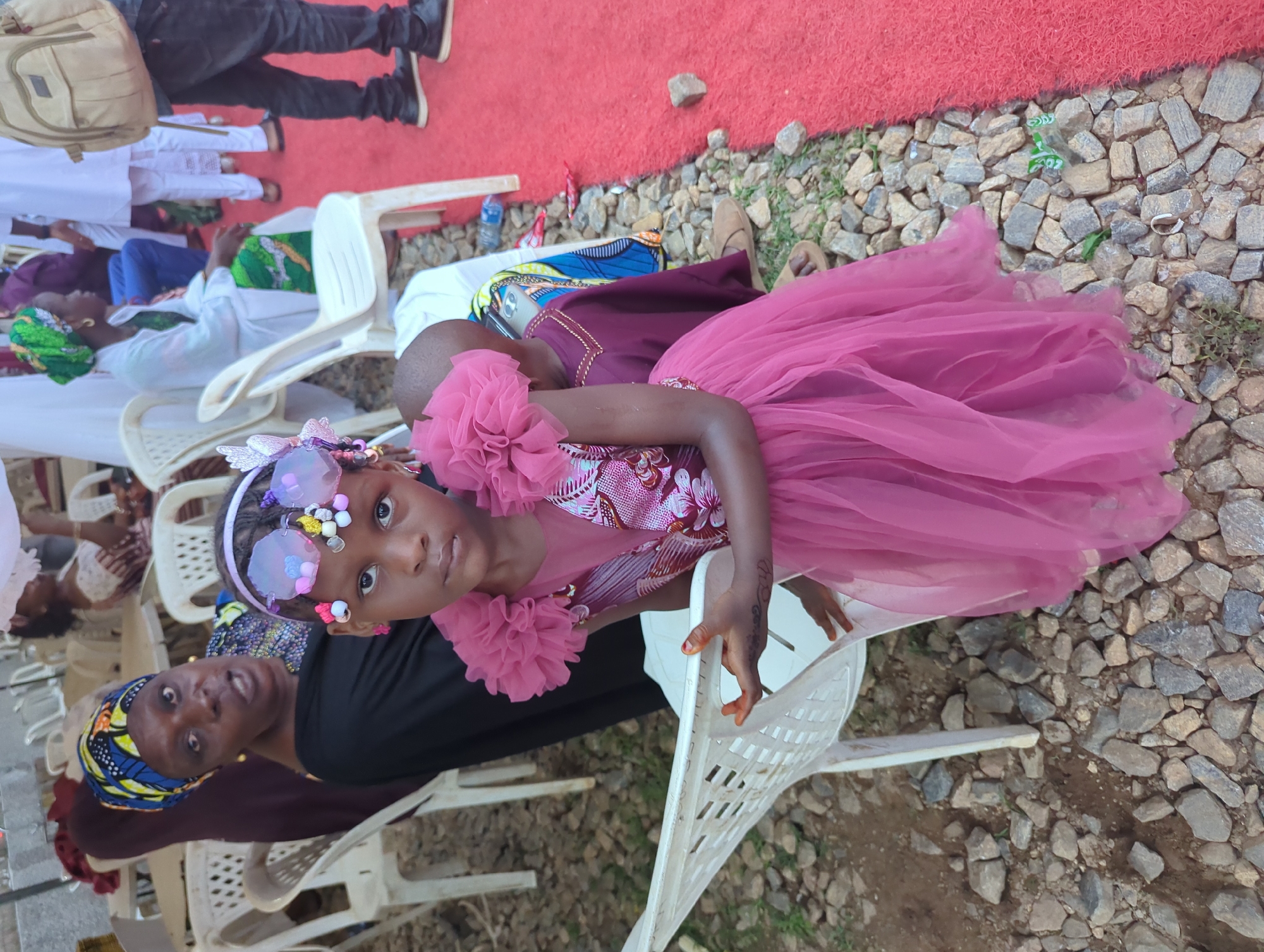 The image captures an outdoor gathering in Kabala, Nigeria, centered on a young girl in the foreground. The girl, estimated to be between four and seven years old, stands with a neutral expression, looking directly at the camera. She wears a sleeveless, two-piece pink/magenta dress; the bodice features a printed fabric with floral or abstract patterns in shades of pink, white, and maroon, while the full skirt is made of layered, sheer pink tulle. Large, ruffled embellishments adorn both shoulders. Her hair is styled with a headband featuring prominent, translucent purple-pink ornaments resembling oversized glasses or butterfly wings, accented with small, colorful beads. Pink stud earrings are visible. Her left hand, displaying reddish staining consistent with henna, rests on the back of a white plastic chair.
Behind and to the left of the girl, an adult woman is seated on a similar white plastic chair. She wears a dark top and a patterned headwrap in blue, yellow, and black fabric. Gold-colored earrings are visible, and her mouth is slightly open, suggesting she is speaking or reacting to something outside the frame. Partially visible behind the main girl, a small child's head, wearing a maroon garment, is present, with a silver mobile phone resting on their lap.
The ground surface is predominantly covered with irregularly shaped small rocks and gravel, interspersed with patches of dry soil and sparse green vegetation. Along the right side of the frame, a bright red, textured outdoor carpet runs parallel to the rocky ground. In the midground and background, multiple white plastic monobloc chairs are scattered, some occupied by blurred figures in various attire, including white garments and headwraps. On the far right, a person's lower body is visible, dressed in dark pants and black shoes, carrying a light brown backpack, standing on the red carpet. A blue plastic water bottle is visible on the ground near the edge of the red carpet. The scene is illuminated by natural outdoor light.