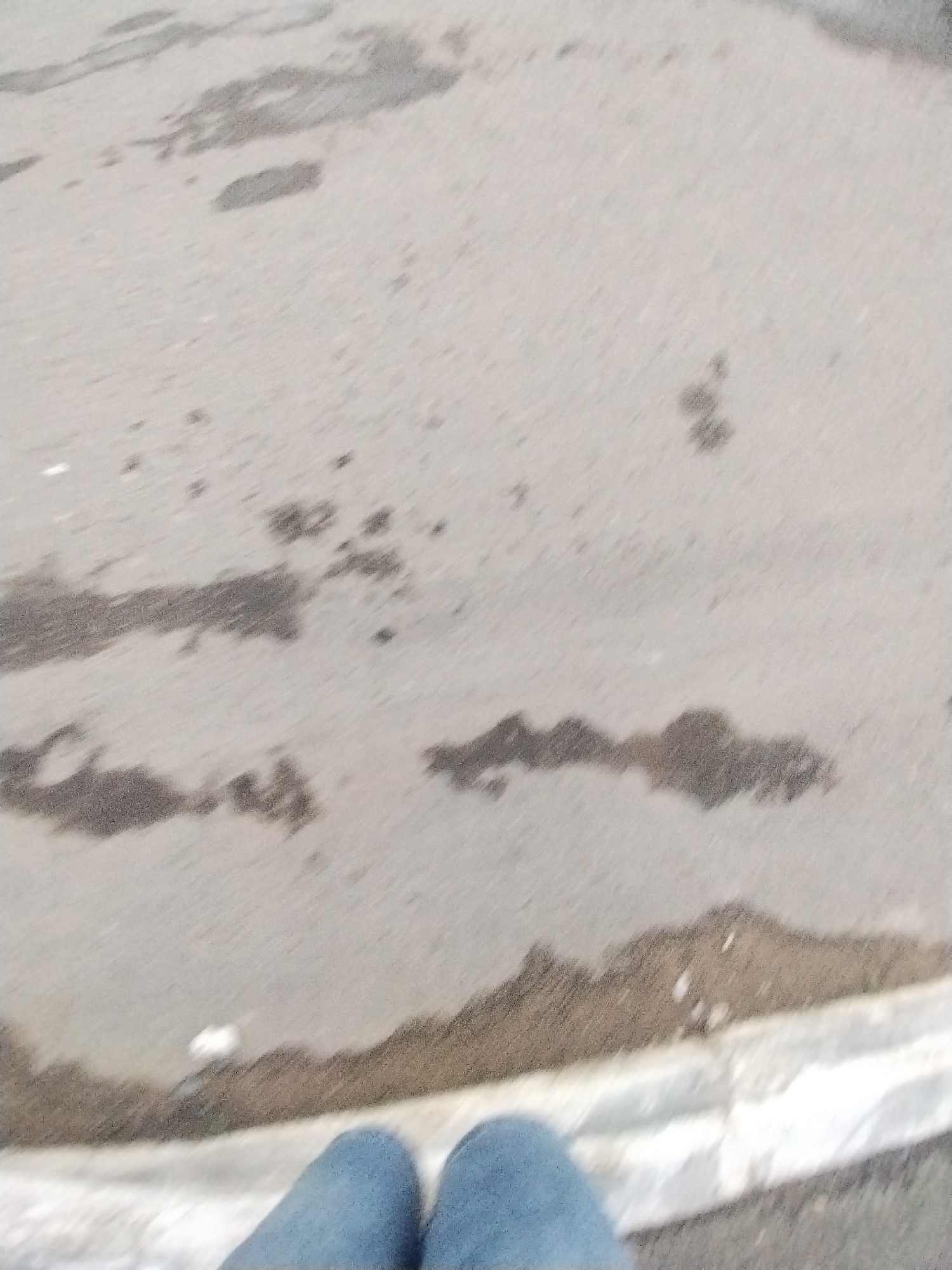 The image shows a close-up, slightly blurry view of a person's legs, clad in blue jeans, standing on an asphalt surface. The asphalt appears wet and discolored in patches, suggesting recent rain or spills. Some small white specks are visible on the ground, possibly litter or debris. The lower portion of the image is dominated by the person's legs and feet, with the ground stretching out ahead. The upper portion of the image is out of focus, showing the wet asphalt pattern. The lighting suggests it might be daytime, but the weather conditions are unclear beyond the presence of moisture on the ground. The scene is neutral and does not convey any specific emotions or a particular event. There is no visible text in the image. The context provided indicates the location is Chaikovsky, Russia.