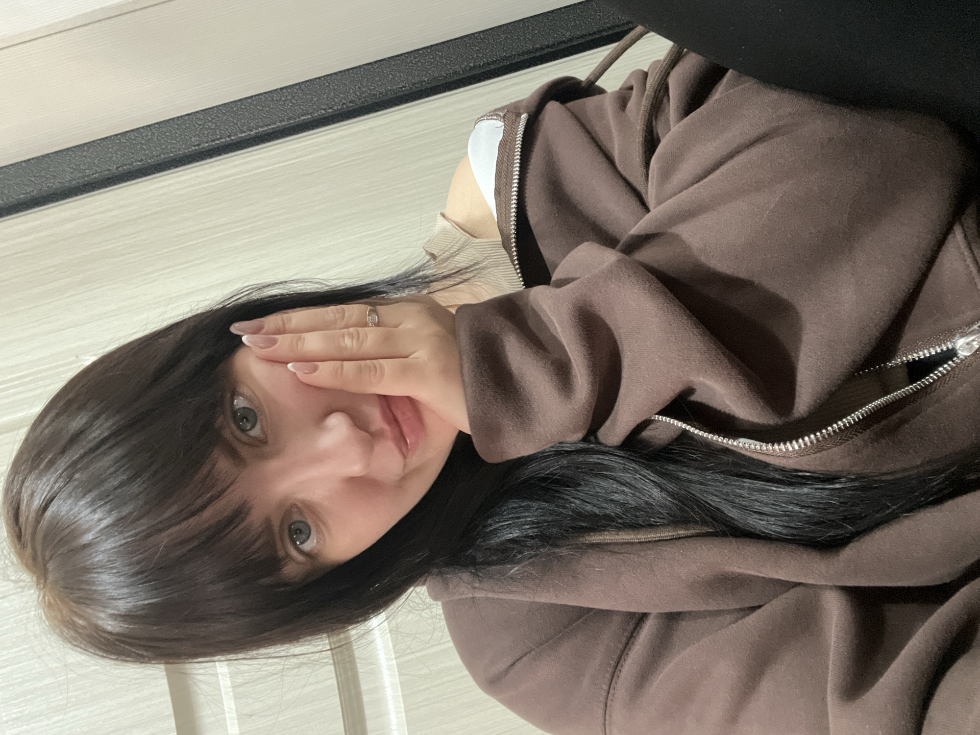A woman with dark hair and light eyes is posing for a selfie in what appears to be an indoor setting. She is wearing a brown zip-up hoodie. Her hand is near her face. The background includes a light-colored wall with a dark trim. The location information is not evident in the picture.