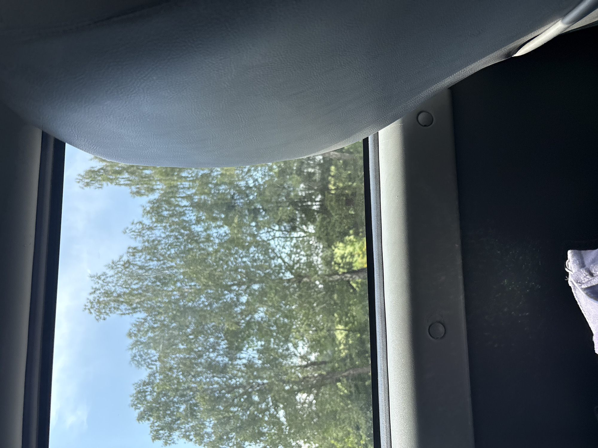 The image is taken from inside a vehicle, looking out a window. In the foreground, on the right side of the image, is the textured, light grey headrest of a vehicle seat. The window shows a bright, sunny day with a clear blue sky and some scattered clouds. Outside the window, there are several tall, leafy green trees, likely in a rural or natural setting. Below the window, within the vehicle, is a dark grey panel with two circular indentations or fixtures visible. At the bottom of the image, a small portion of a folded, light blue fabric item is visible, possibly clothing. The scene appears to be a snapshot of a journey through a green, possibly forested, area. This image does not contain any elements that indicate a specific location in the city of Мастоцкі сельскі Савет, Belarus, or any other specific city.