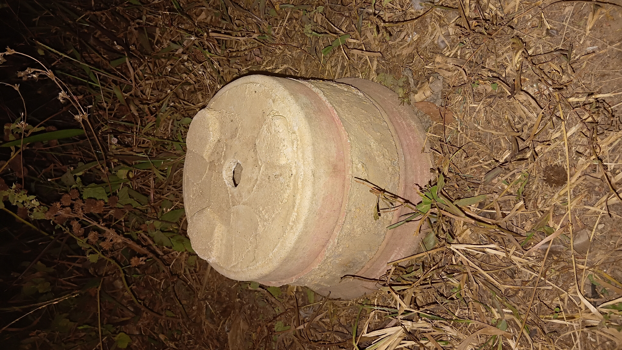 The image shows a circular, layered object made of concrete or stone, placed on the ground amidst dry vegetation and some green plants. The object appears to be a repurposed pot or base, with a hole in the center of its top surface. The texture of the object is rough and weathered, with some pinkish discoloration on the lower layers. The background consists of dark foliage and dry grass, suggesting an outdoor setting, possibly in a neglected or natural area. The lighting suggests it was taken at night or in low-light conditions. The location is specified as Prabumulih, Indonesia, which could imply an urban or suburban environment with pockets of unkempt natural growth.