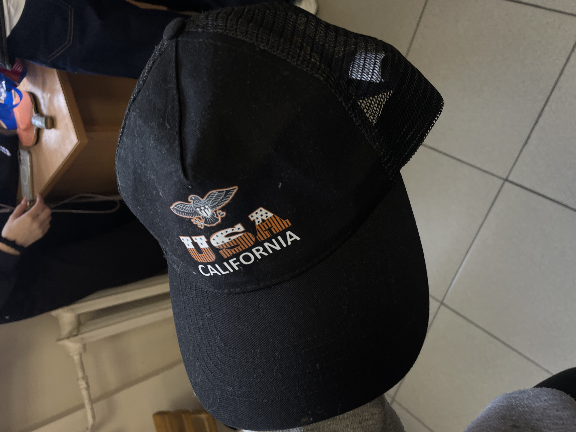 An overhead, slightly angled photograph captures a black trucker cap with a mesh back, held by a person whose gray-sleeved arm is partially visible. The cap features an embroidered design on its front panel. An orange and white eagle emblem is positioned above the word "USA," where the letters themselves are patterned with orange and white stripes and stars, evoking the American flag. Below this, the word "CALIFORNIA" is stitched in white capital letters. The cap's fabric appears to be black canvas or cotton for the front and brim, with a synthetic mesh for the back panels, and shows some light dust or lint.
In the background, a warm-toned wooden table or desk is visible, featuring various indistinct objects including a blue item and an orange item. A hand, possibly belonging to another individual, holds a smartphone next to this table. Further in the background, a person wearing blue jeans is partially seen. The floor consists of light-colored, possibly off-white, square tiles with dark grout lines. A white, decorative architectural element, resembling a railing or radiator cover, is also present. The scene is set indoors, suggesting a casual environment in Novoe Devyatkino, Russia. The lighting is diffused, consistent with an indoor setting.
