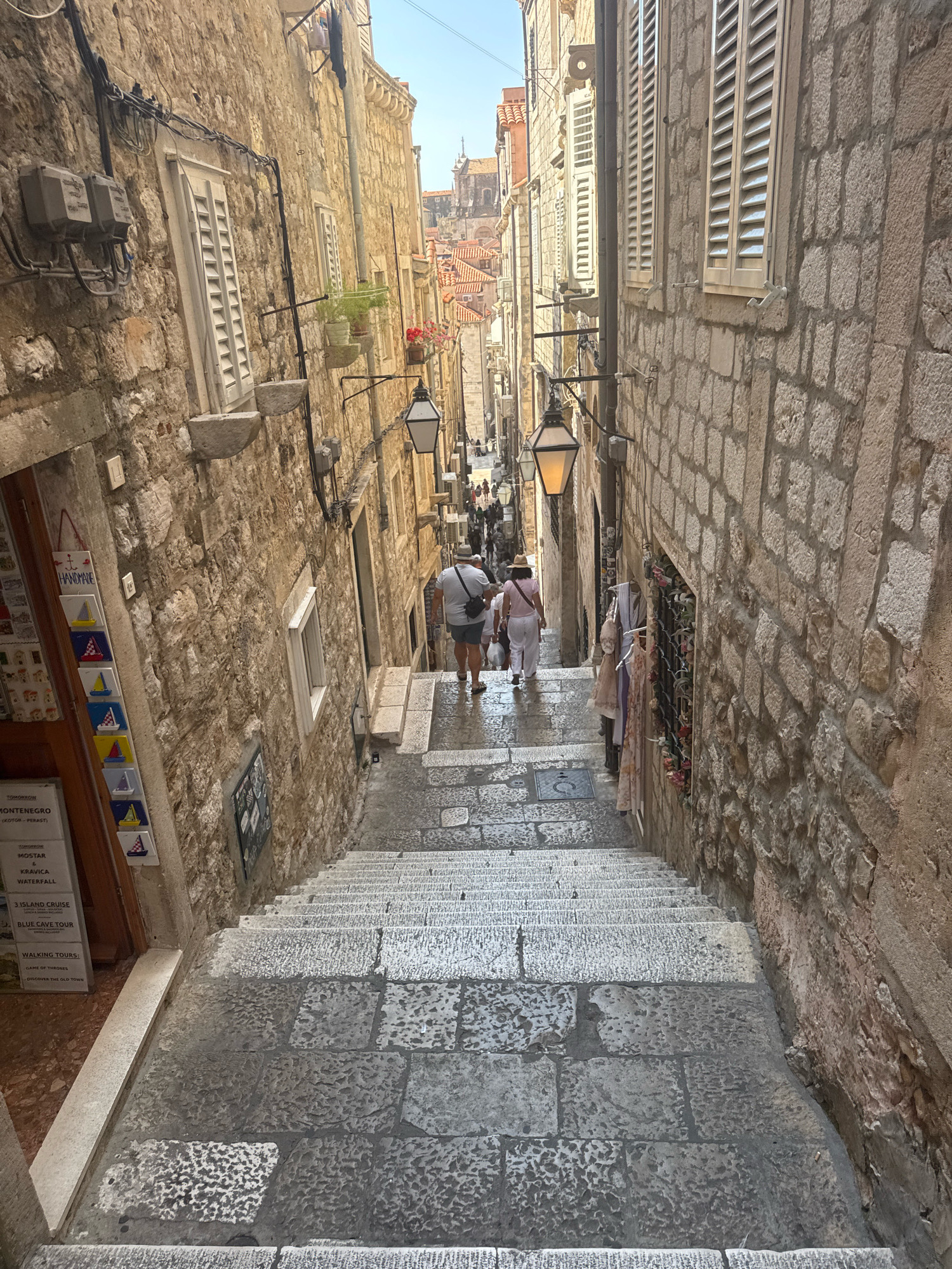 This image depicts a steep stone staircase descending through a narrow, historic street in Dubrovnik, Croatia. The stone buildings on either side are ancient and weathered, with characteristic stone facades and wooden shutters. There are visible electrical conduits and boxes running along the walls, indicating the age and adaptation of the buildings. Traditional street lamps are mounted on the walls.
On the left side of the staircase, a shop is visible with various souvenirs displayed. These include small ceramic tiles with nautical motifs and posters advertising tours such as "Montenegro," "Mostar Waterfall," and "Game of Thrones."
Two individuals are walking down the stairs in the middle ground. The person in front, facing away from the camera, wears a white hat, a pink top, and white pants. The person behind them, also facing away, wears a light-colored t-shirt and shorts, with a black bag slung across their chest. Their feet are wet, suggesting the stairs may be damp or recently cleaned. Further down the staircase, other pedestrians are visible, blurred by distance.
The overall impression is of a quintessential Dubrovnik street scene, characterized by its ancient architecture, vibrant tourist activity, and the unique charm of its stone passageways. The lighting suggests it is daytime, with the sun casting shadows on the stairs.