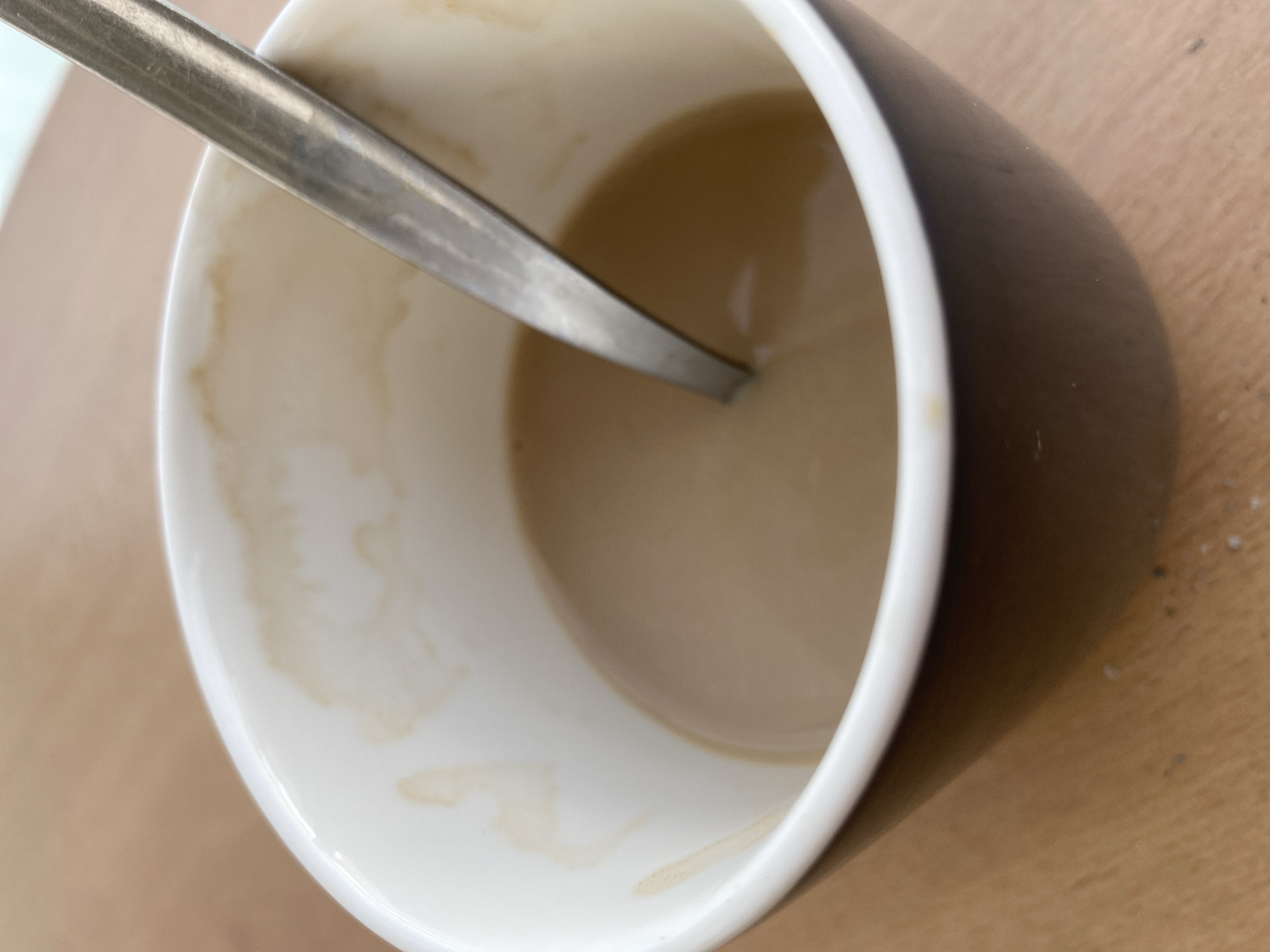 A close-up, overhead shot captures a dark mug filled with a light brown liquid, presumably coffee with milk. A silver spoon rests inside, submerged in the liquid, its handle angled towards the upper right of the frame. The inner rim of the mug displays faint, dried brown stains, suggesting previous use. The mug sits on a light brown, wooden surface, possibly a table. The overall scene is domestic and unremarkable.