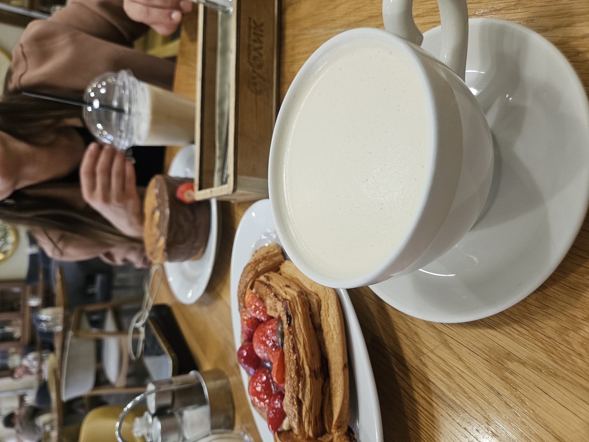 The image, taken from a slightly elevated perspective and rotated 90 degrees clockwise, showcases a table laden with food and drinks in what appears to be a cafe or restaurant setting. The scene is primarily focused on various delectable items arranged on a light-colored wooden table.
In the prominent right foreground, a white ceramic cup holds a creamy, light-colored beverage, likely a frothy coffee drink such as a latte or cappuccino, resting on a matching saucer. Adjacent to it, in the lower center, a golden-brown, flaky pastry, possibly a fruit tart or Danish, is presented on a rectangular white plate. This pastry is generously adorned with glistening red fruits, which appear to be sliced strawberries or cherries.
In the mid-ground, a person with long brown hair and wearing a brown top is partially visible on the left, holding a tall, clear plastic cup with a black straw. This cup contains a light brown beverage, possibly an iced coffee or smoothie. Behind this individual, a round, dark-colored cake, likely chocolate, is placed on a small white plate.
A notable object in the upper-middle is a wooden box-like structure or display, featuring text in Cyrillic script. The visible text spells out "БУБЛИК" (BUBLIK), which is a Russian or Ukrainian term for a type of bread roll or bagel. In the lower left corner, a silver and clear condiment dispenser, possibly a sugar shaker, is partially visible. The background is softly blurred, indicating other elements of the cafe environment, such as tables or chairs. The overall ambiance suggests a relaxed and enjoyable moment, typical of a cafe outing.