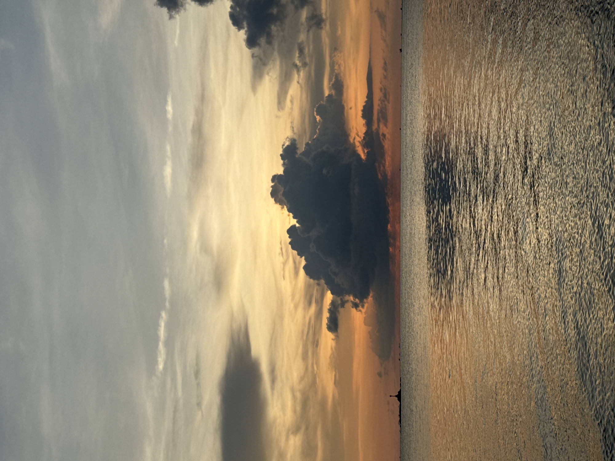 The image captures a serene sunset over the water in Baan Sri Thanu, Thailand. The sky is filled with varying shades of clouds, ranging from light gray to dark, ominous formations. The sun casts a golden and orange glow across the sky, reflected on the rippled water surface. A few indistinct dark shapes are visible along the horizon, possibly structures or boats. The overall atmosphere is tranquil.
