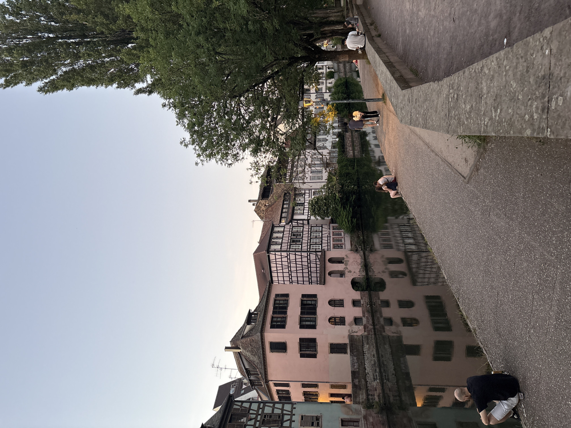 The image captures a scene in Strasbourg, France, likely along a canal in the Petite France district, renowned for its picturesque half-timbered houses. Several such buildings line the waterway, their reflections visible in the water. A tree's foliage partially obscures the buildings. Along the canal-side pavement, multiple people are present. One individual is crouched near the water's edge, while a couple is standing further down, and another person sits by a tree in the distance. The sky is a pale blue, suggesting either early morning or late evening. A concrete wall defines the edge of the canal. The image is composed with a slight rotation, tilting the scene somewhat.