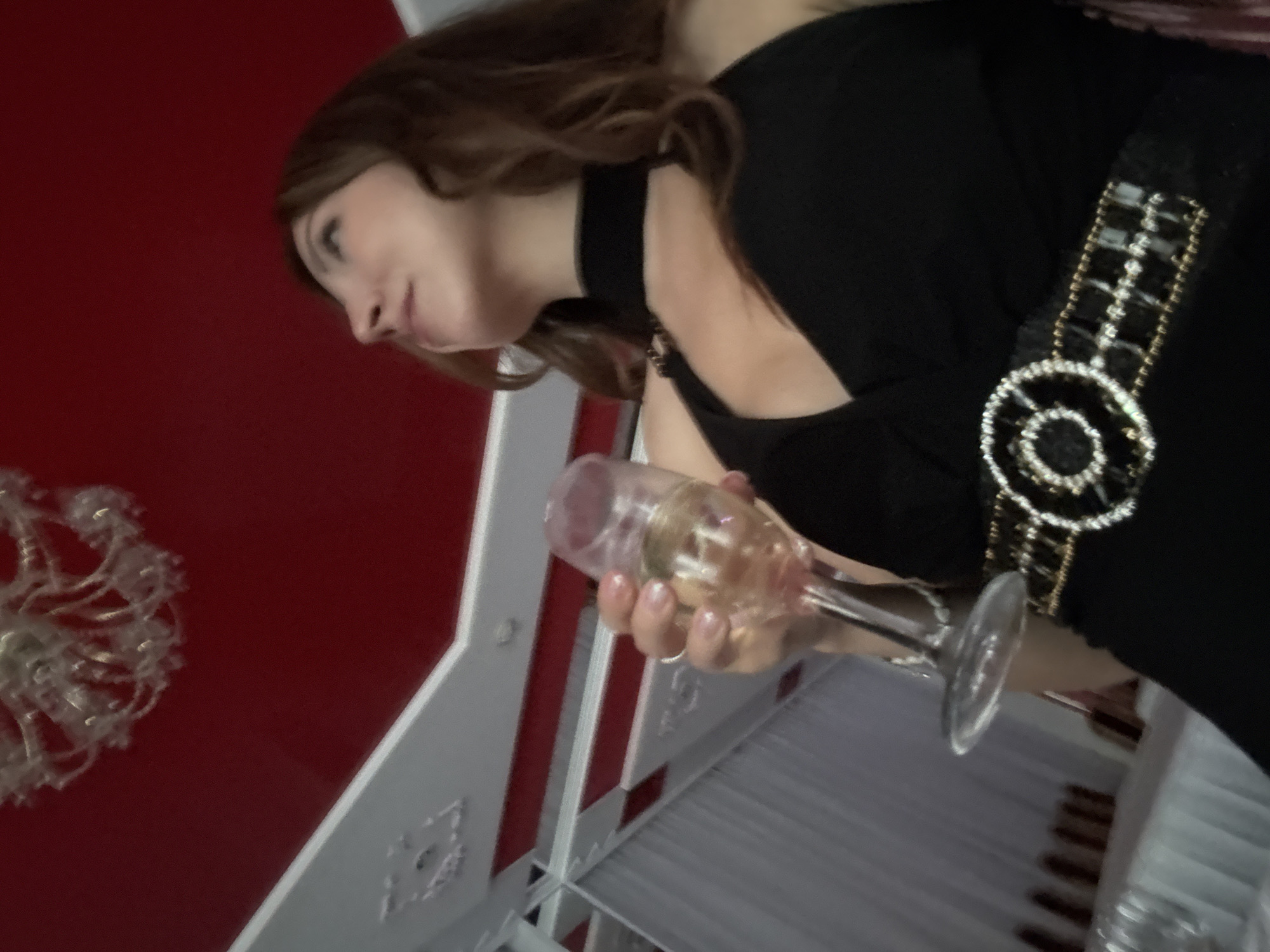 A medium-shot, low-angle, indoor, slightly blurred, full-color photograph shows a woman holding a champagne flute in her right hand. The woman has shoulder-length brown hair and is wearing a black, sleeveless dress with a cut-out neckline and a decorative belt. The belt is black with gold trim and circular embellishments made of white stones. The woman is looking upwards and to the right. The background consists of a red ceiling with decorative white molding and a chandelier. A white curtain and tables are visible in the lower left corner. The location is specified as Kryvyi Rih, Ukraine.