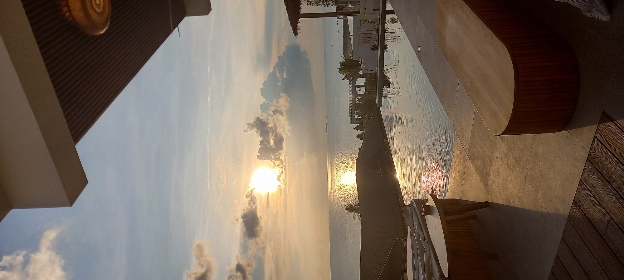 The image shows a sunset view from what appears to be a luxury residence in Baan Wok Tum, Thailand. The sun is positioned in the lower-center of the frame, partially obscured by clouds, and casting golden reflections on the water surface. A long, narrow swimming pool stretches towards the sea, reflecting the buildings and foliage. Palm trees and modern architectural structures are visible along the pool's edge. A portion of a wooden bench and some deck furniture are in the foreground on a tiled patio area.