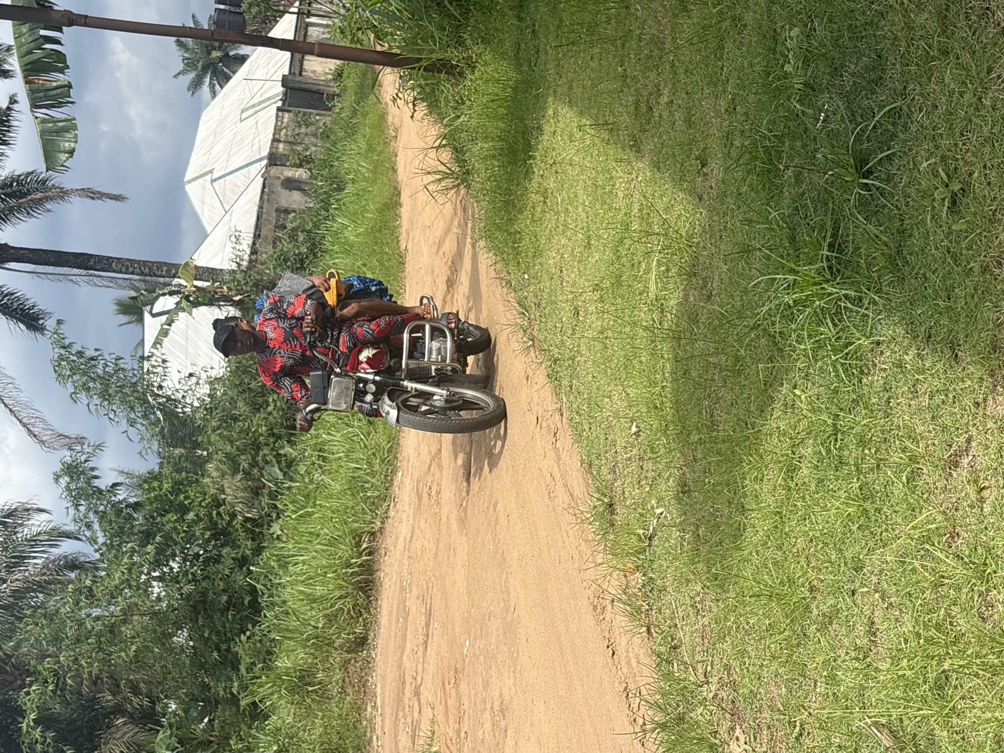 A person is riding a motorcycle down a dirt road in Ikot Ekpene, Nigeria. The rider is wearing a red and black patterned shirt. A second person is sitting behind the driver, with both feet on the motorcycle foot rests. The road is bordered by green grass on both sides, and there is lush vegetation to the left of the road, including a palm tree. In the background, there is a light-colored building with a white roof under a cloudy sky. A wooden pole is visible near the top-left of the image.