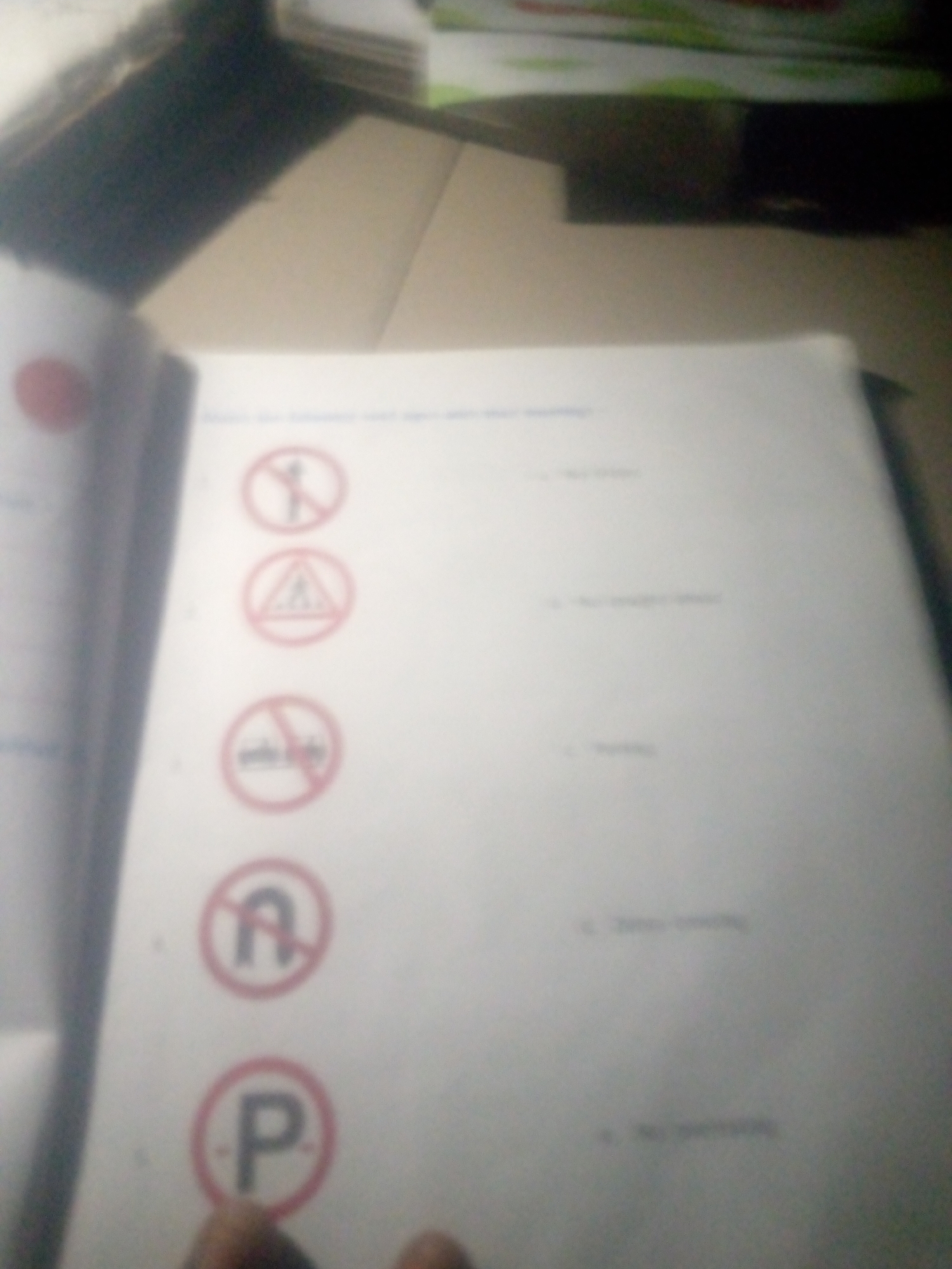 The image shows a page of a workbook or test with several traffic signs and corresponding multiple-choice answers. The workbook is open, with the left page partially visible, showing a red circular symbol. The right page is the main focus, displaying five traffic signs arranged vertically.
The first sign is a circular red sign with a diagonal red line across it, enclosing a black silhouette of a person walking. The corresponding answer is "a. two wheelers".
The second sign is a triangular red warning sign with a black border, containing a black silhouette of a person walking across a striped rectangle, indicating a pedestrian crossing. The corresponding answer is "b. No straight ahead".
The third sign is a circular red sign with a diagonal red line across it, enclosing a symbol that appears to be a car. The corresponding answer is "c. No U-turn".
The fourth sign is a circular red sign with a diagonal red line across it, enclosing a black letter "A". The corresponding answer is "d. Zebra crossing".
The fifth sign is a circular red sign with a black letter "P" inside. The corresponding answer is "e. No parking".
The background is blurred but appears to be a surface with some books or papers stacked. The lighting is dim, suggesting it might be indoors. The location is not discernible from the image.