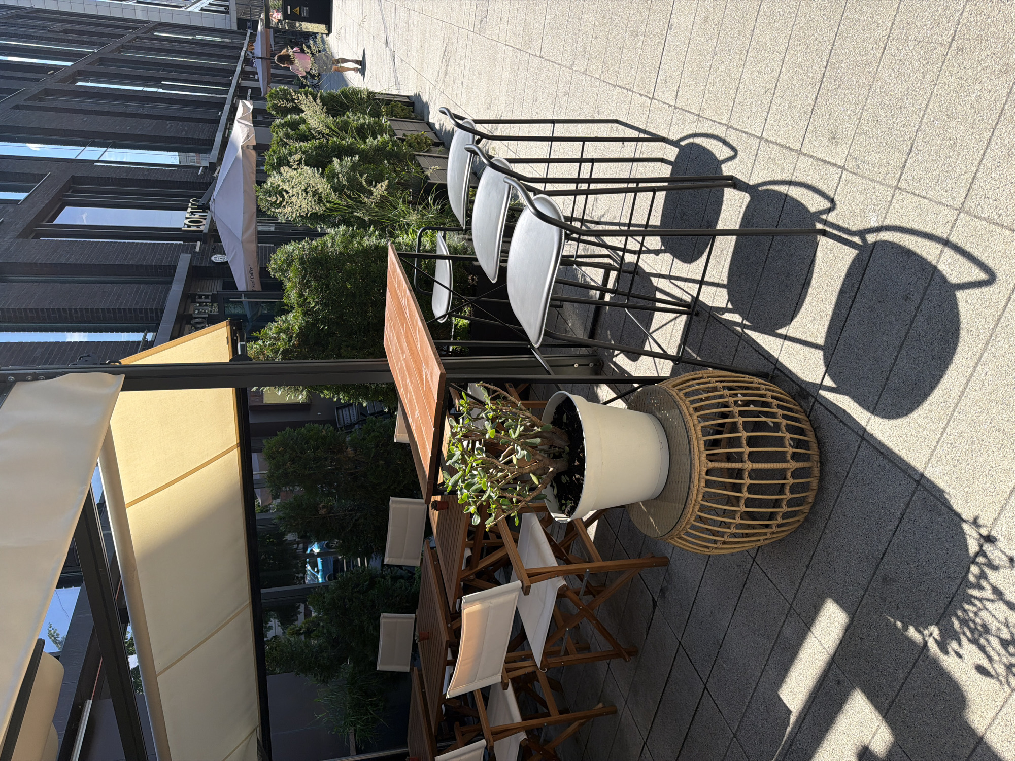 The image depicts an outdoor seating area of a commercial establishment, possibly a cafe or restaurant, located on a city sidewalk. In the foreground, several wooden tables and chairs are arranged, with some tables covered by a beige awning. A potted plant sits on a round, wicker stool. To the right, a row of three high-top stools with gray seats and black metal frames is positioned next to a wooden table. The ground is paved with light-colored tiles. In the background, a modern, dark brick building with large windows dominates the scene, with trees and umbrellas visible along the sidewalk. A person is walking away from the camera on the sidewalk in the distance. The lighting suggests it is daytime, with shadows cast on the ground by the tables, chairs, and plants.