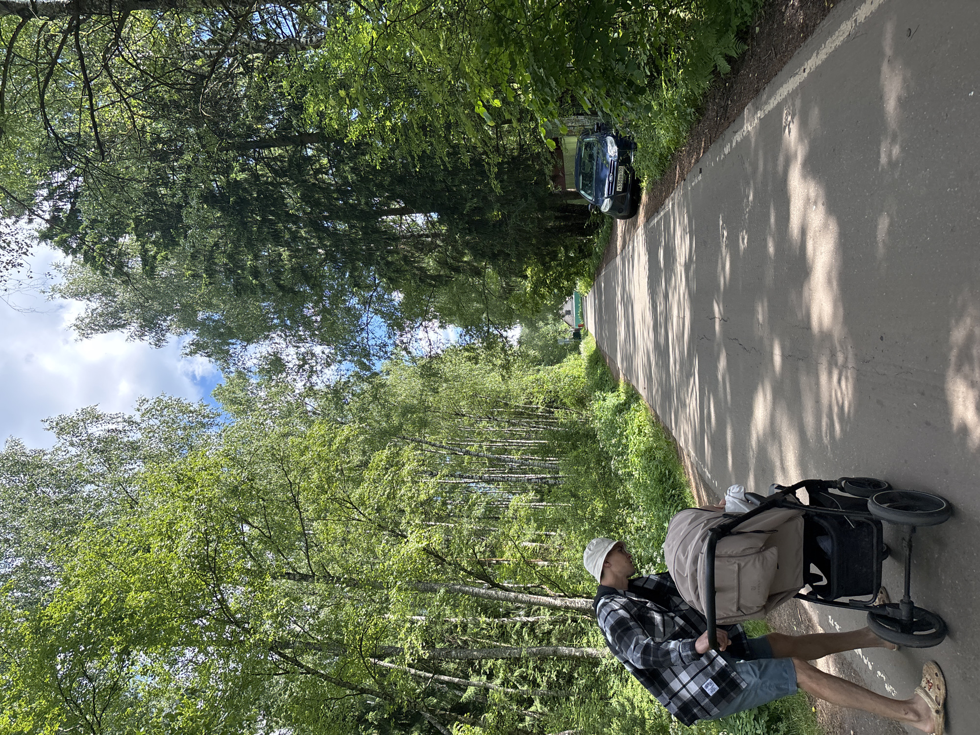 In Мещёры, Russia, a person wearing a plaid shirt, blue shorts, and a white bucket hat pushes a stroller along a paved road lined with trees. The stroller appears to be beige and black, and it is occupied by a baby or young child. A dark blue car is parked on the right side of the road, partially obscured by foliage. The road is surrounded by lush green trees, and the sky is blue with some clouds. The sun is casting shadows on the road, indicating it is daytime. The scene appears to be a peaceful, outdoor setting in a wooded area.