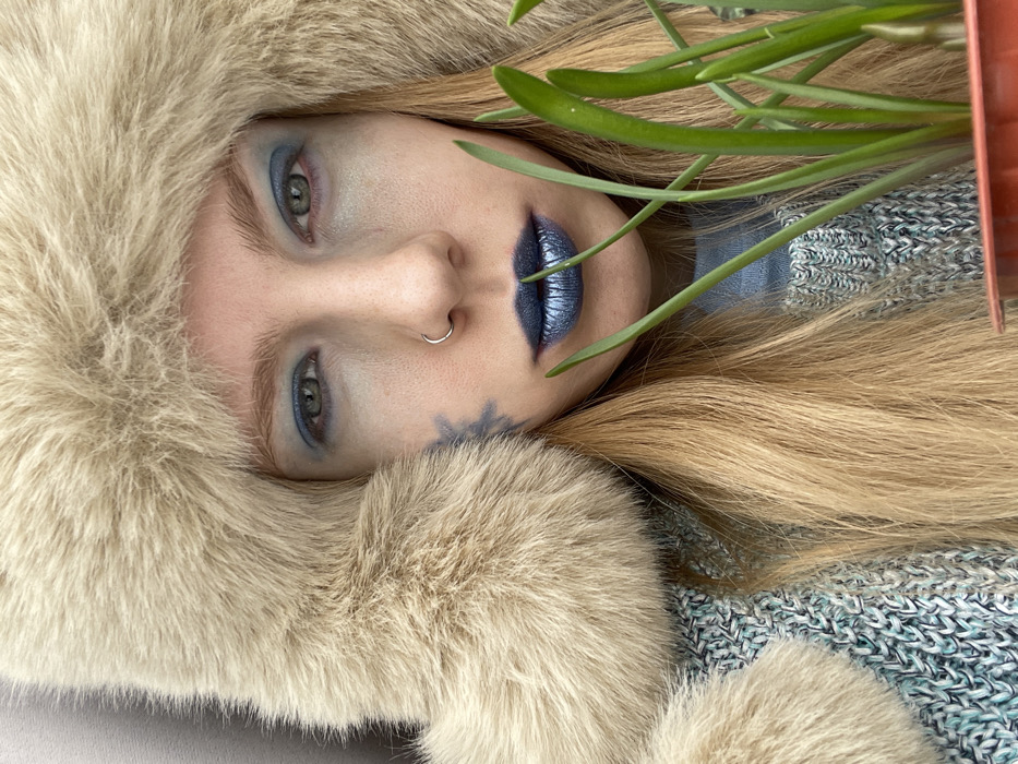 A close-up portrait depicts a single individual, appearing to be a young adult female, against an indistinct, light-grey background. The image is taken indoors or in a sheltered outdoor setting; no specific location within Izhevsk, Russia, is discernible. The individual has long, straight blonde hair and light-colored eyes. They are wearing a light-colored (tan/beige) fur trapper hat and a knitted sweater composed of light blue, white, and grey threads, with a plain light blue garment visible beneath. Their face features heavy blue eyeshadow on the eyelids and metallic dark blue lipstick. A silver hoop nose ring is visible in the left nostril. A blue, snowflake or star-shaped design is painted on the left cheek, below the eye. The individual looks directly into the camera with a neutral expression. In the foreground, positioned to the lower right, a terracotta-colored pot holds several green, leafy plants, which partially obscure the individual's lower face and right shoulder.