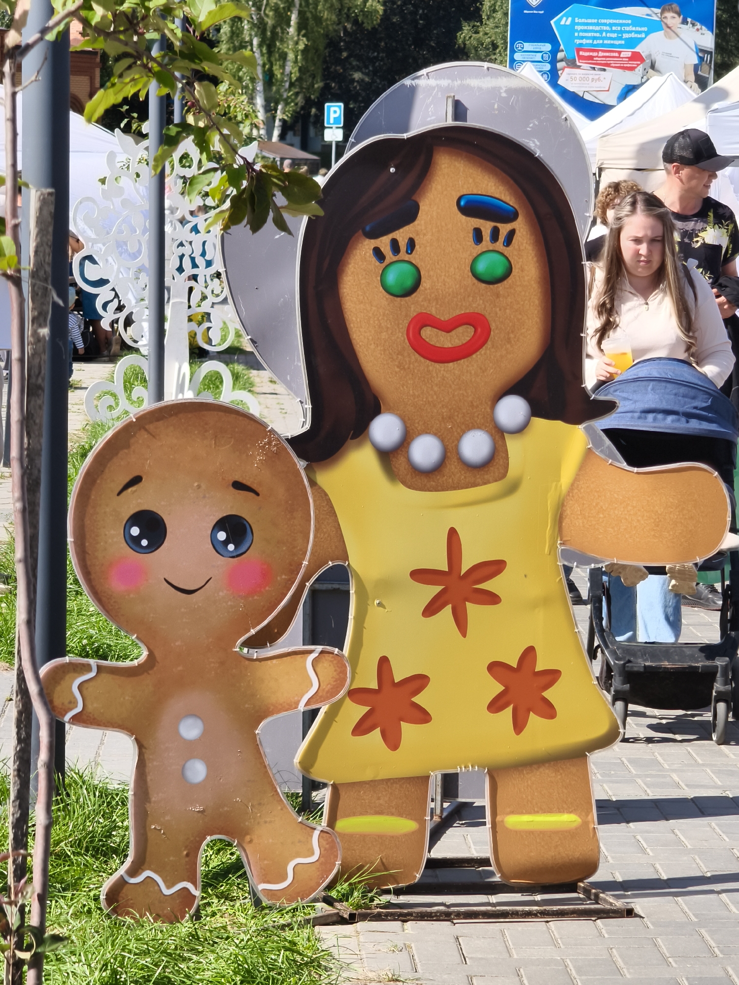 The image captures a sunny outdoor scene featuring a large, cutout display of two gingerbread people, one adult-sized and one child-sized, in Chaikovsky, Russia. The adult gingerbread figure is dressed in a yellow dress adorned with orange flower shapes and a pearl necklace. The child gingerbread figure stands to the left of the adult figure. In the background, a woman with light brown hair is visible, holding a beverage and looking down. Behind her, a man wearing a black baseball cap and a patterned t-shirt is seen walking. Tents and a sign with Russian text are also present in the background, suggesting a market or event. The foreground consists of green grass and a paved walkway. The overall atmosphere appears to be festive and casual.